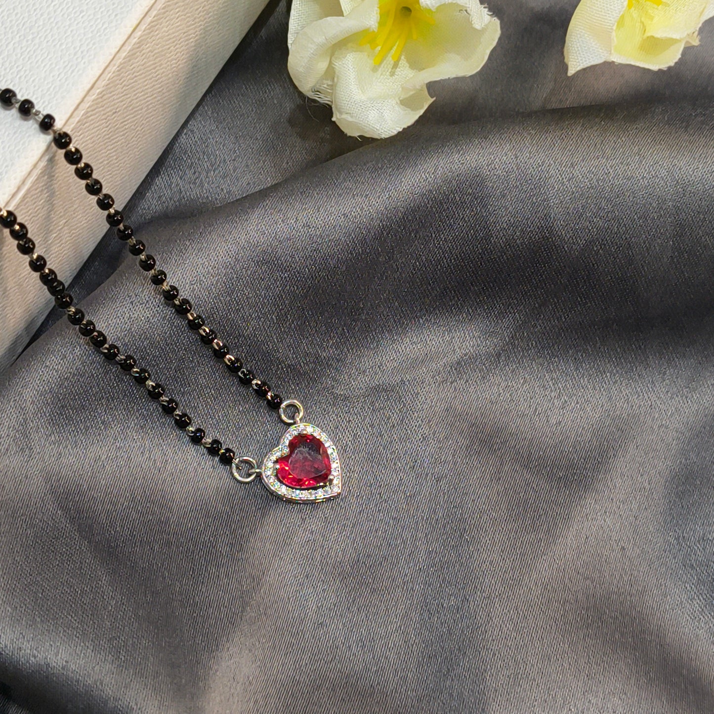 Necklace with a heart-shaped red gemstone pendant on a gray fabric background with white flowers.