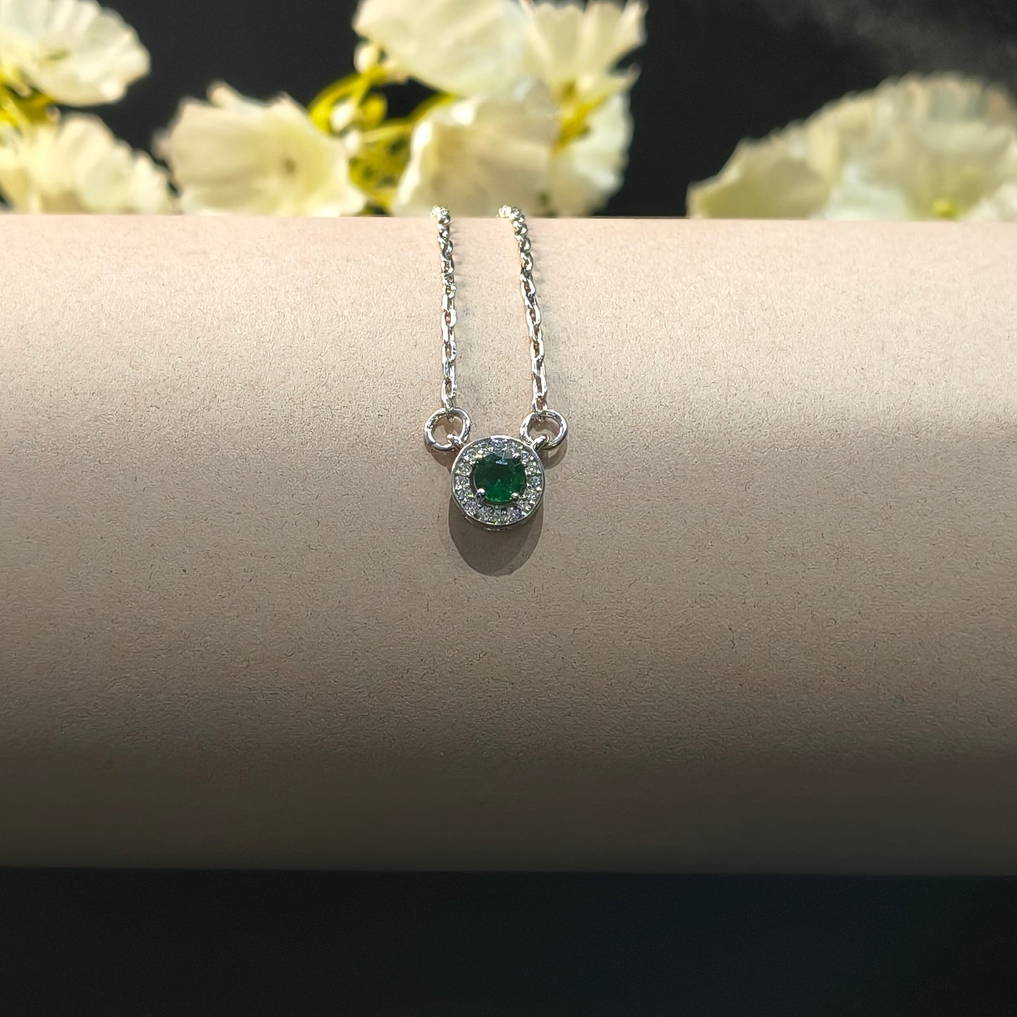 Silver necklace with a green gemstone pendant on a beige stand with white flowers in the background