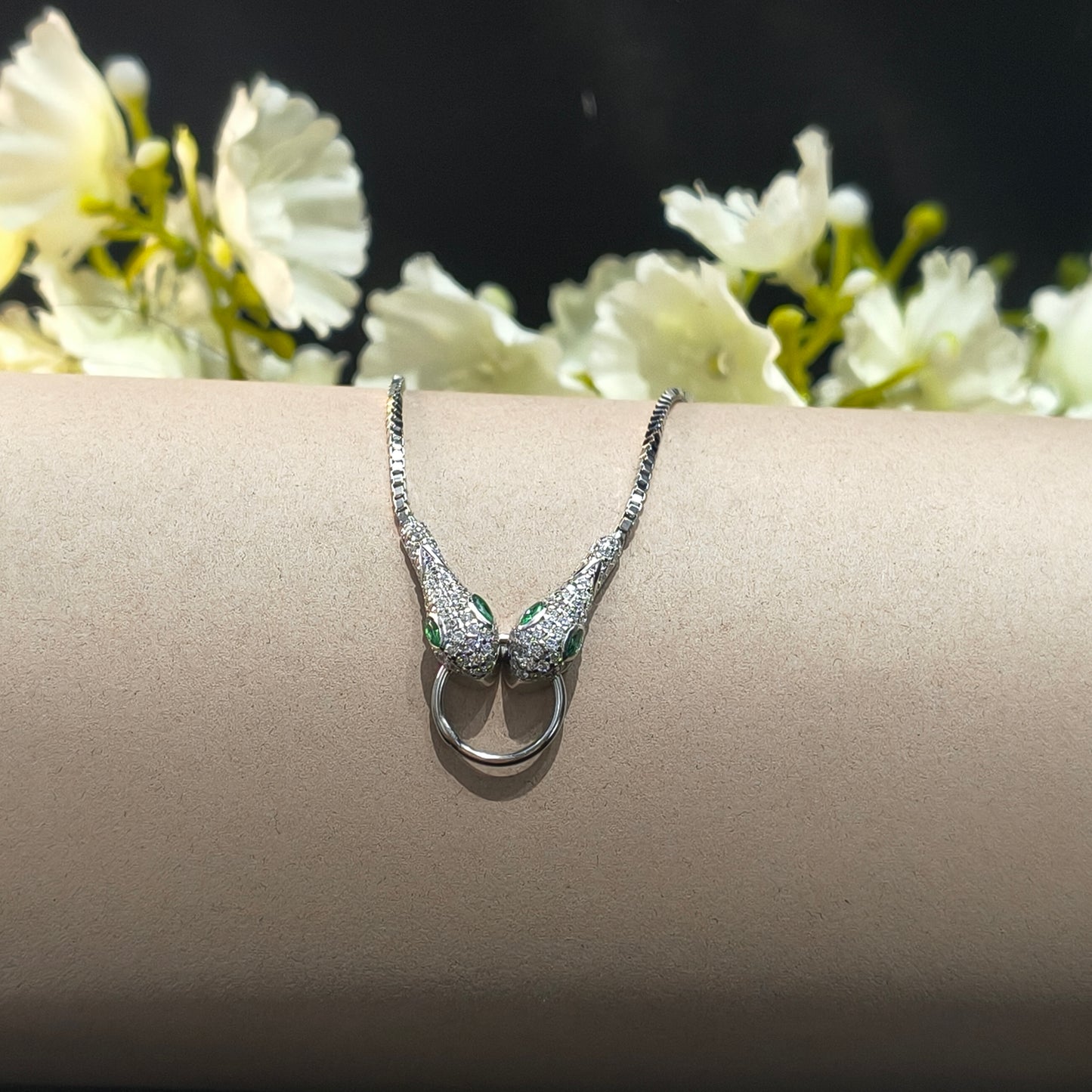 Silver bracelet with green stones on a beige stand with white flowers in the background