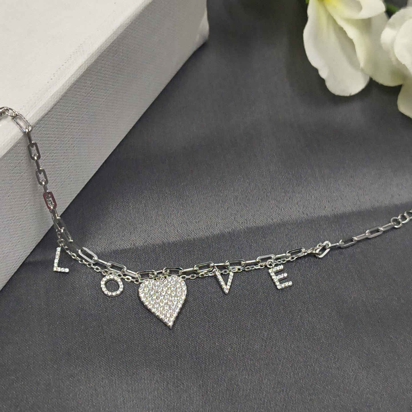 Silver bracelet with heart charm and 'LOVE' letters on a gray surface with white flowers.
