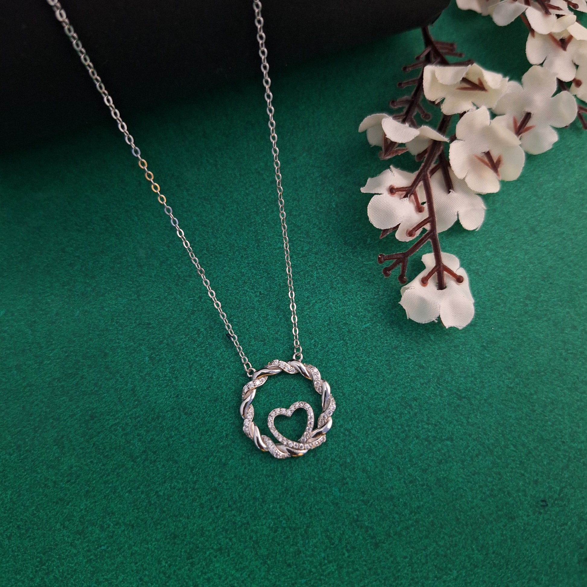 Silver necklace with a heart-shaped pendant on a green background with white flowers.