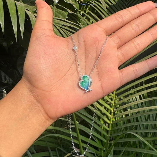 Hand holding a necklace with a green and blue butterfly pendant against a green leafy background