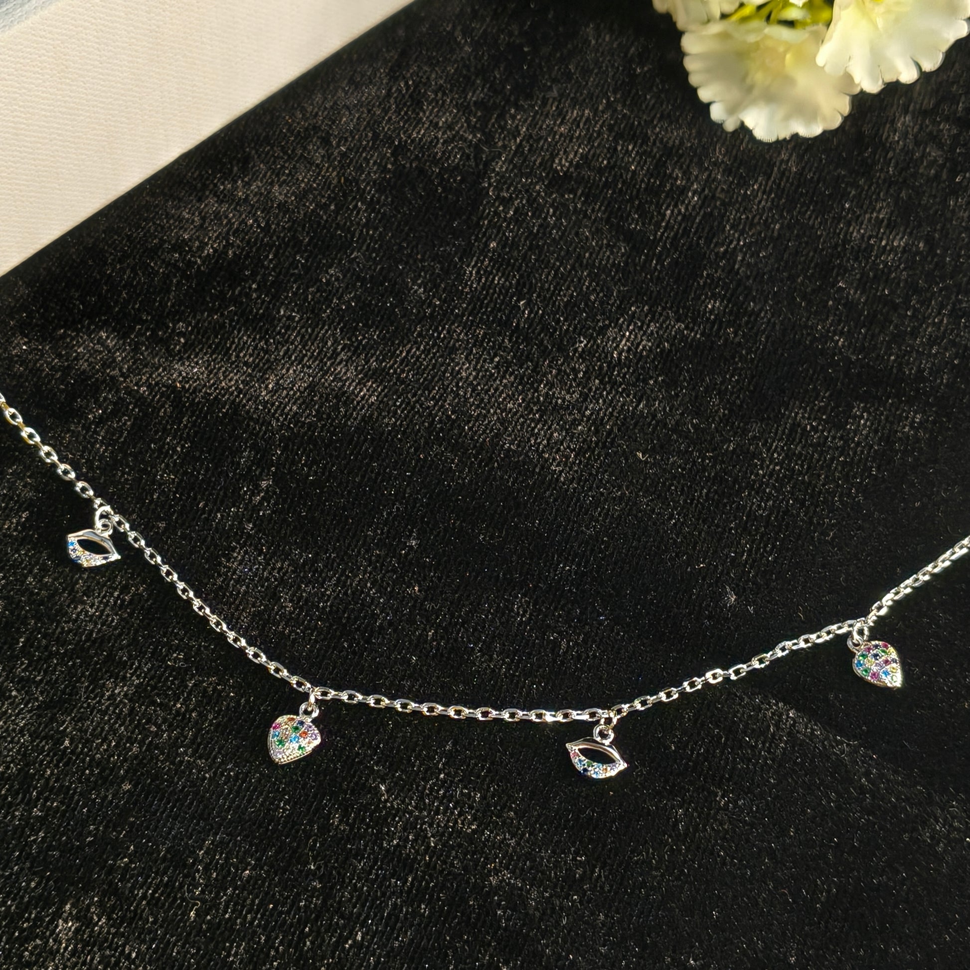 Delicate silver necklace with heart-shaped charms on a black surface with white flowers.