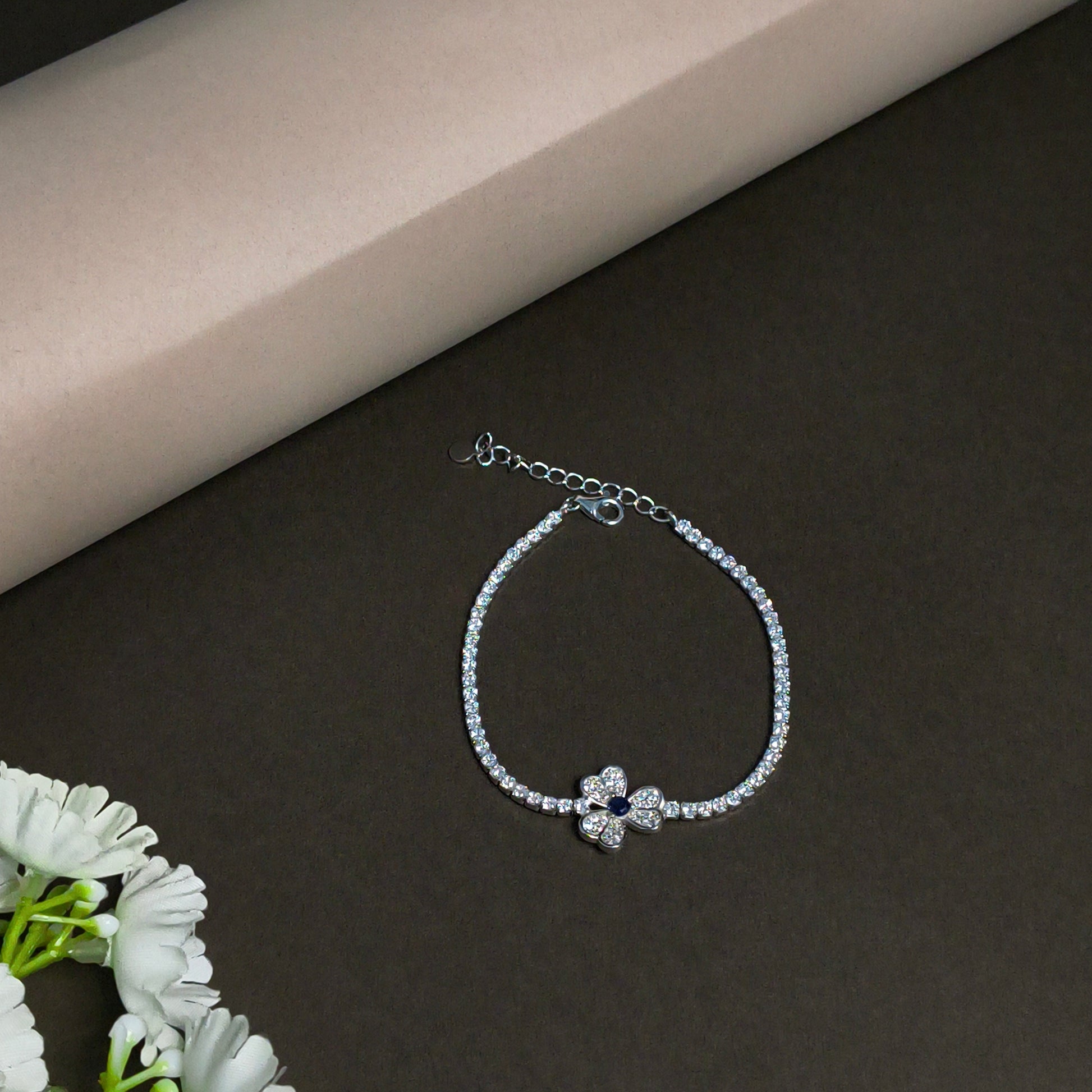 Silver bracelet with a flower-shaped clasp on a dark surface with a beige corner and white flowers.