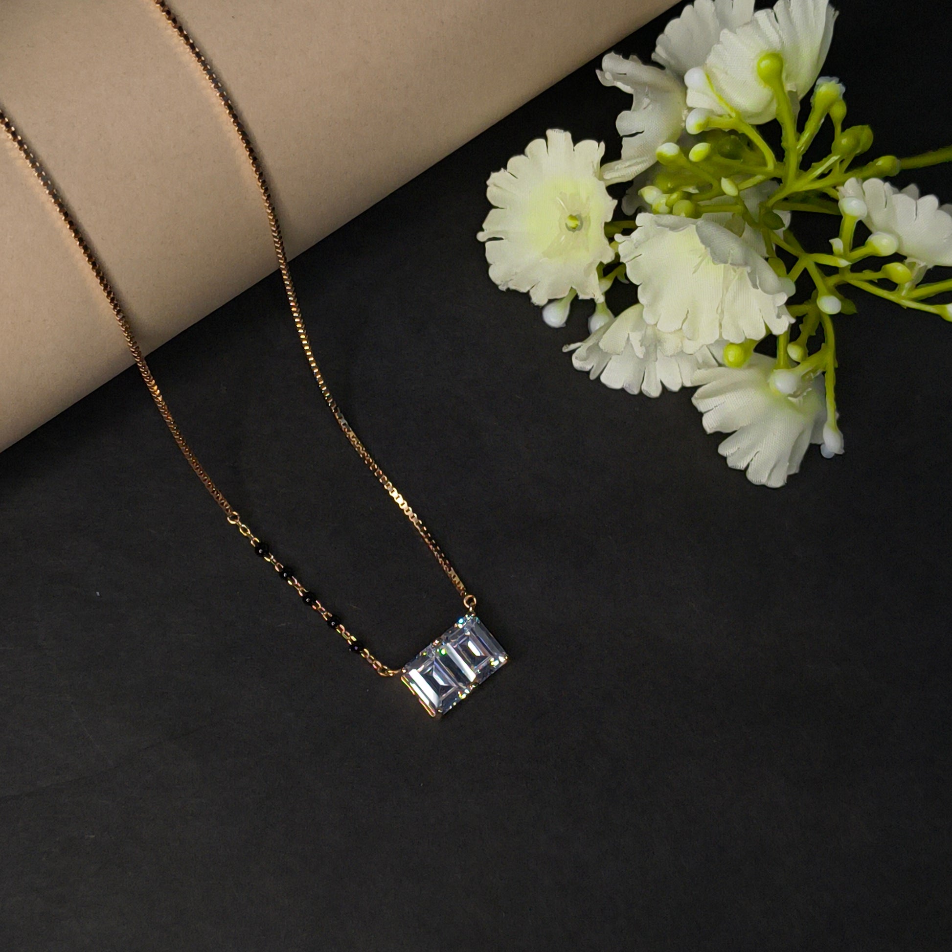 Gold necklace with a rectangular pendant on a dark surface with white flowers.