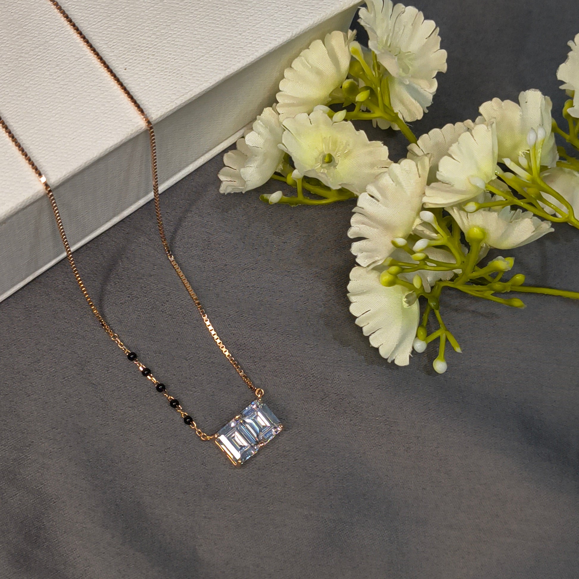 Gold necklace with a rectangular pendant on a gray surface with white flowers.