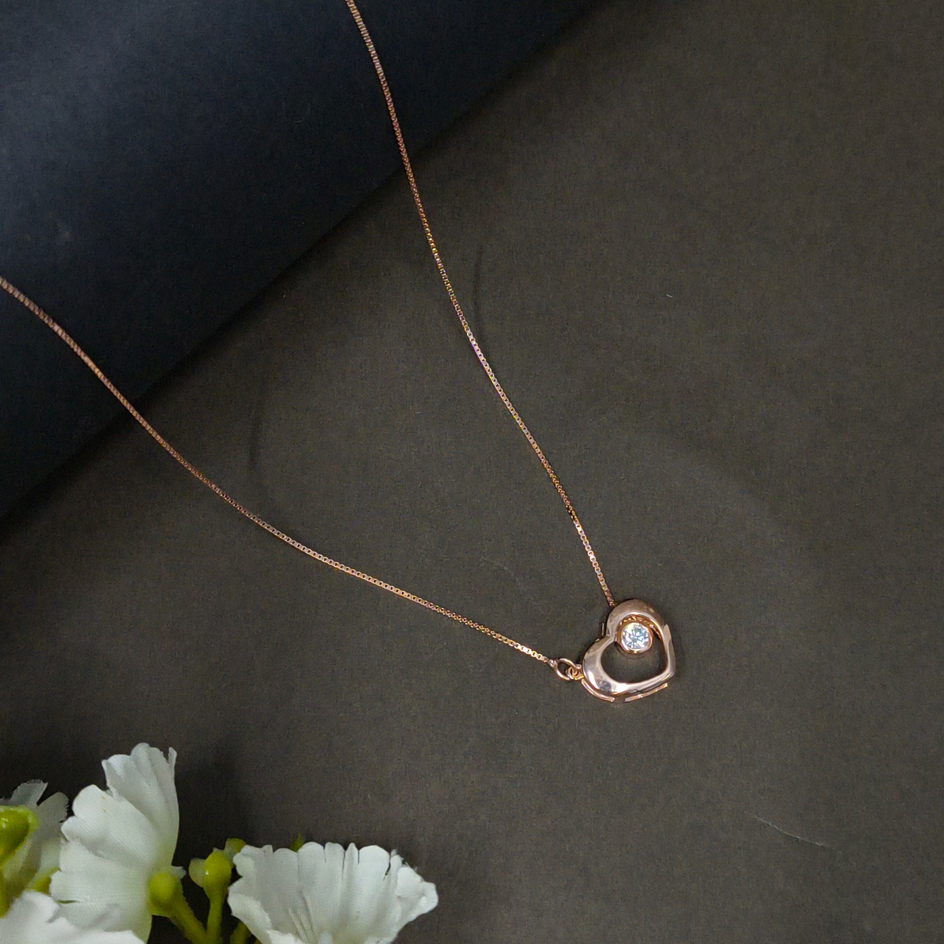 Rose gold necklace with a heart-shaped pendant on a dark background with white flowers.