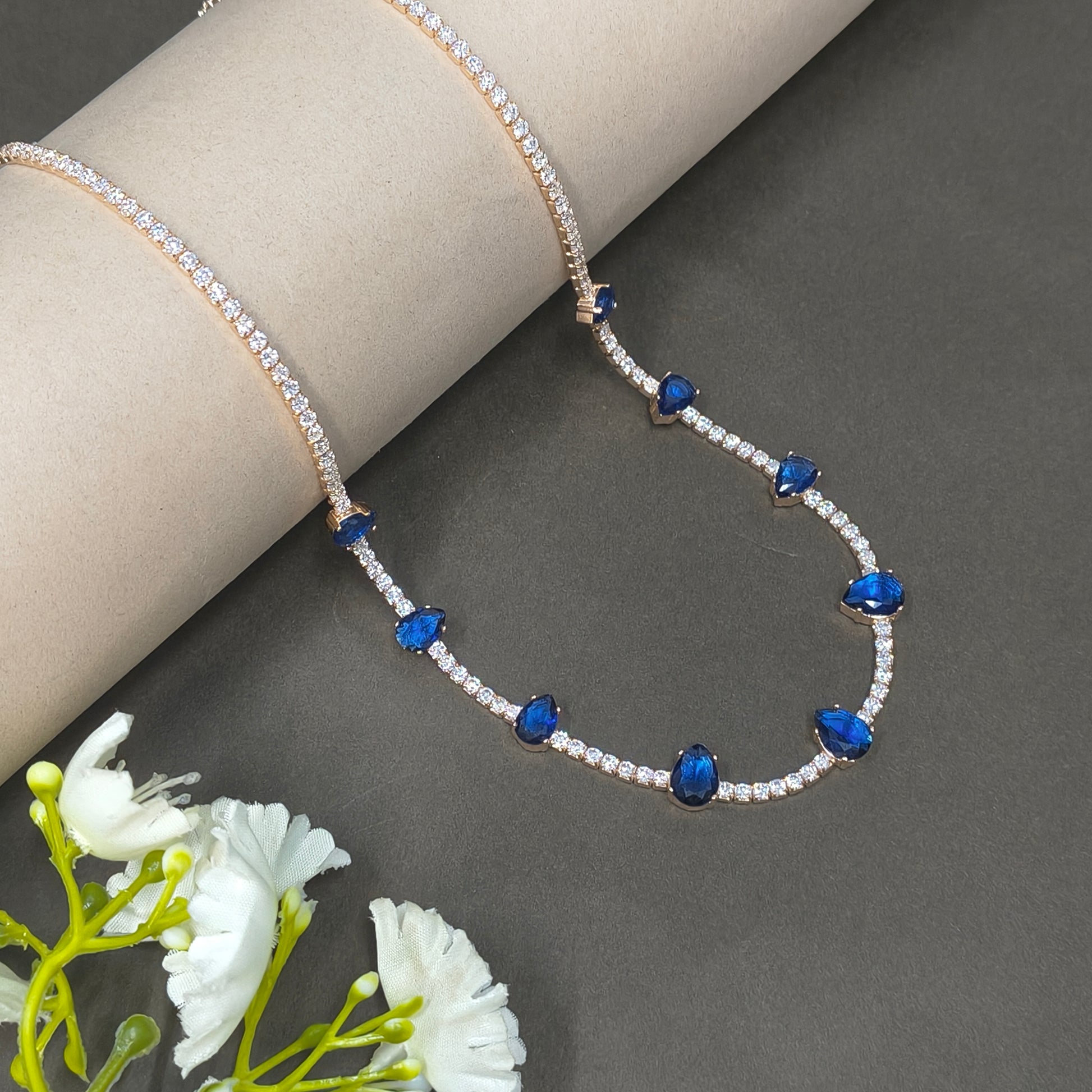Heart-shaped necklace with blue and clear stones on a gray background with white flowers.