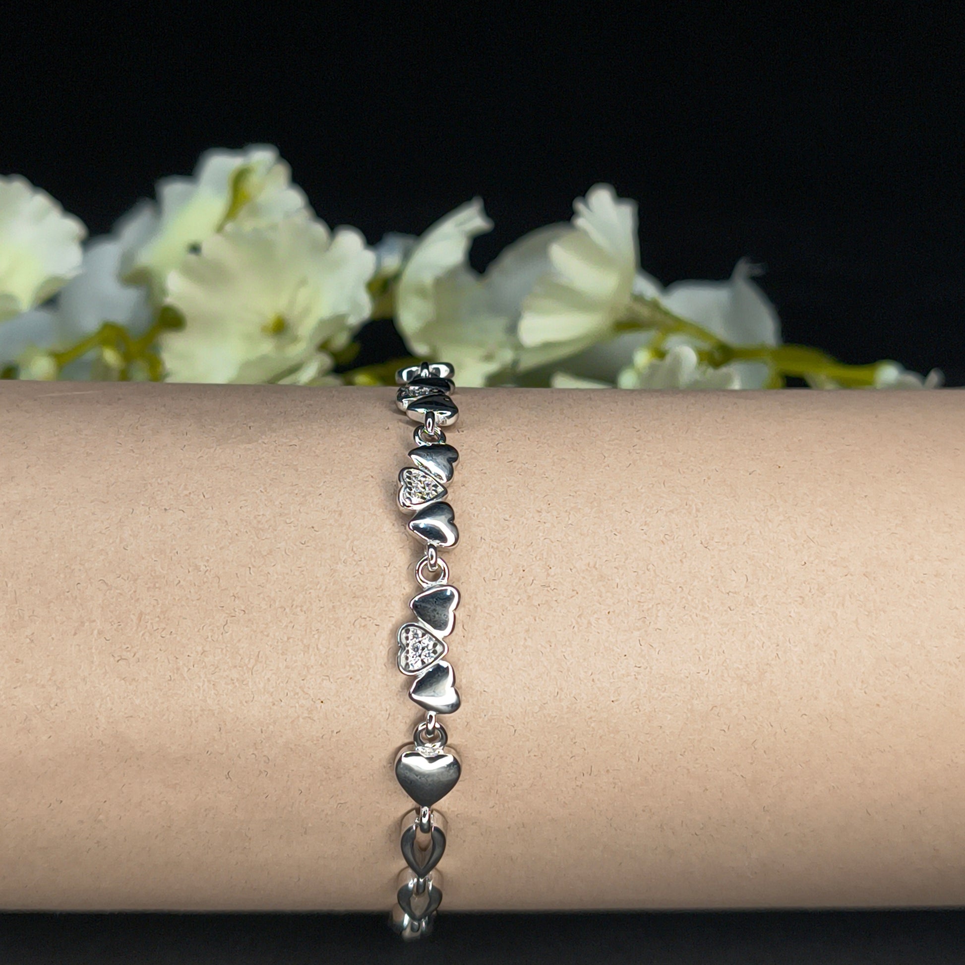 Silver bracelet with heart-shaped charms on a wrist against a dark background with white flowers.