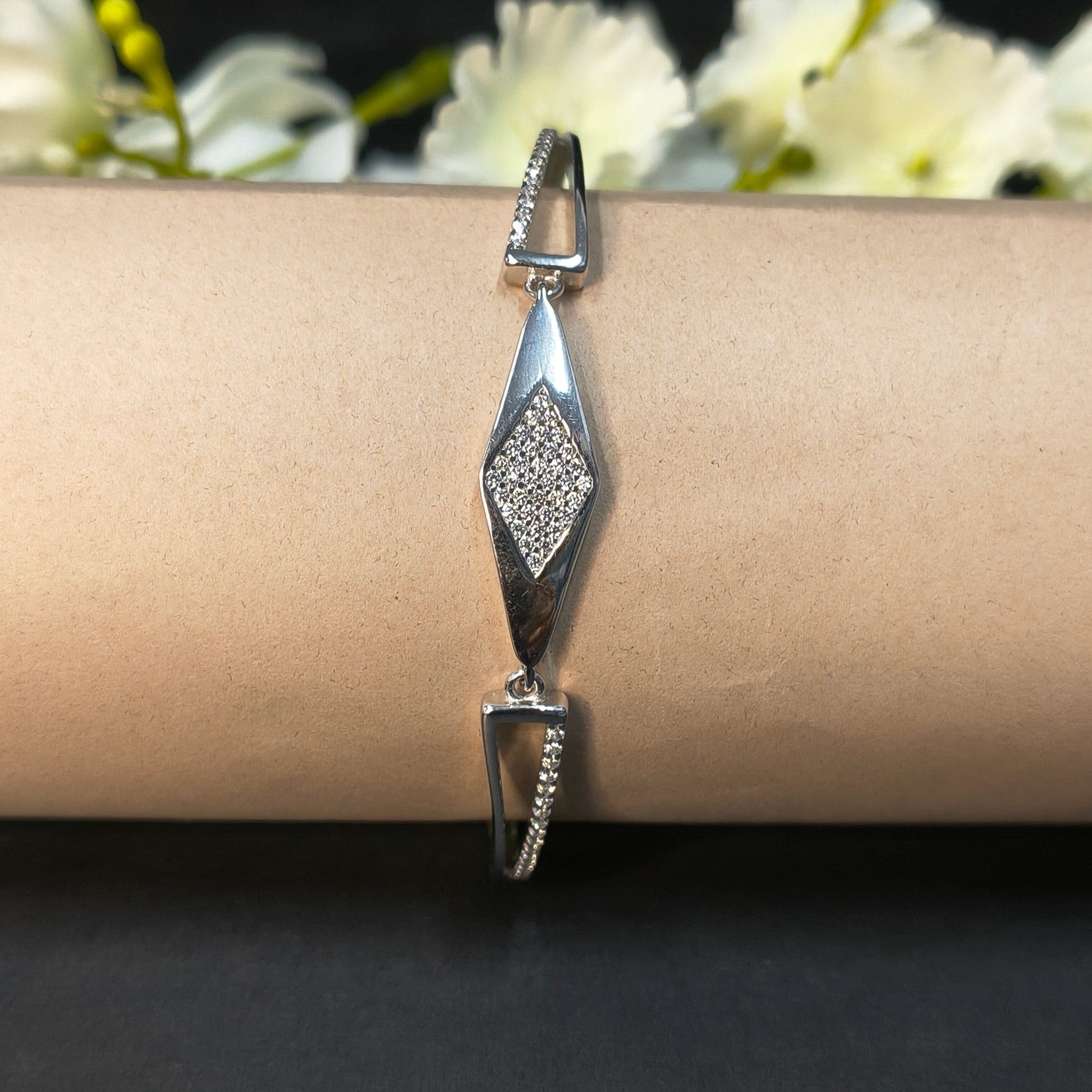 Silver bracelet with diamond-shaped design on a wrist against a dark background with white flowers.