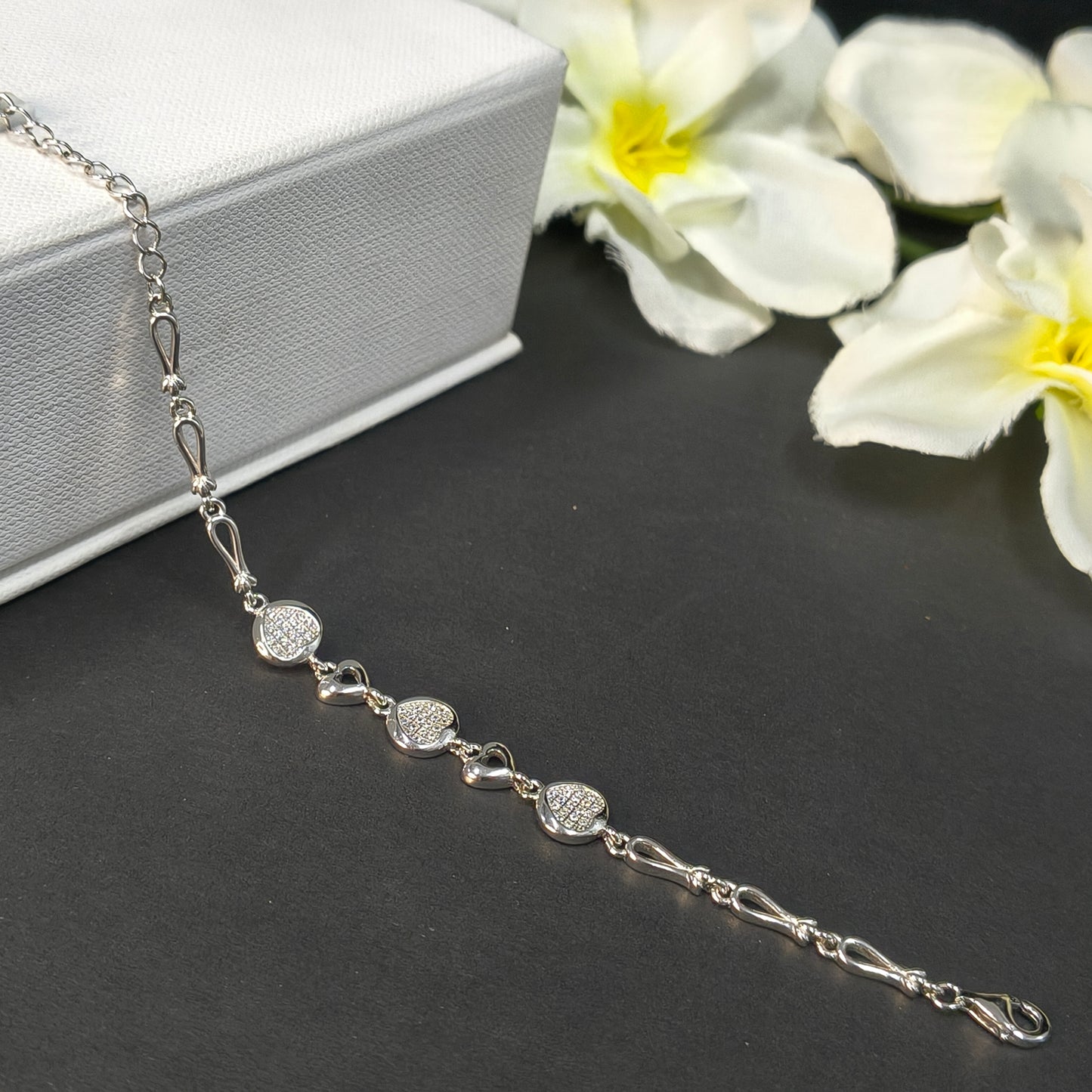 Silver bracelet with heart-shaped charms on a dark surface with white flowers.