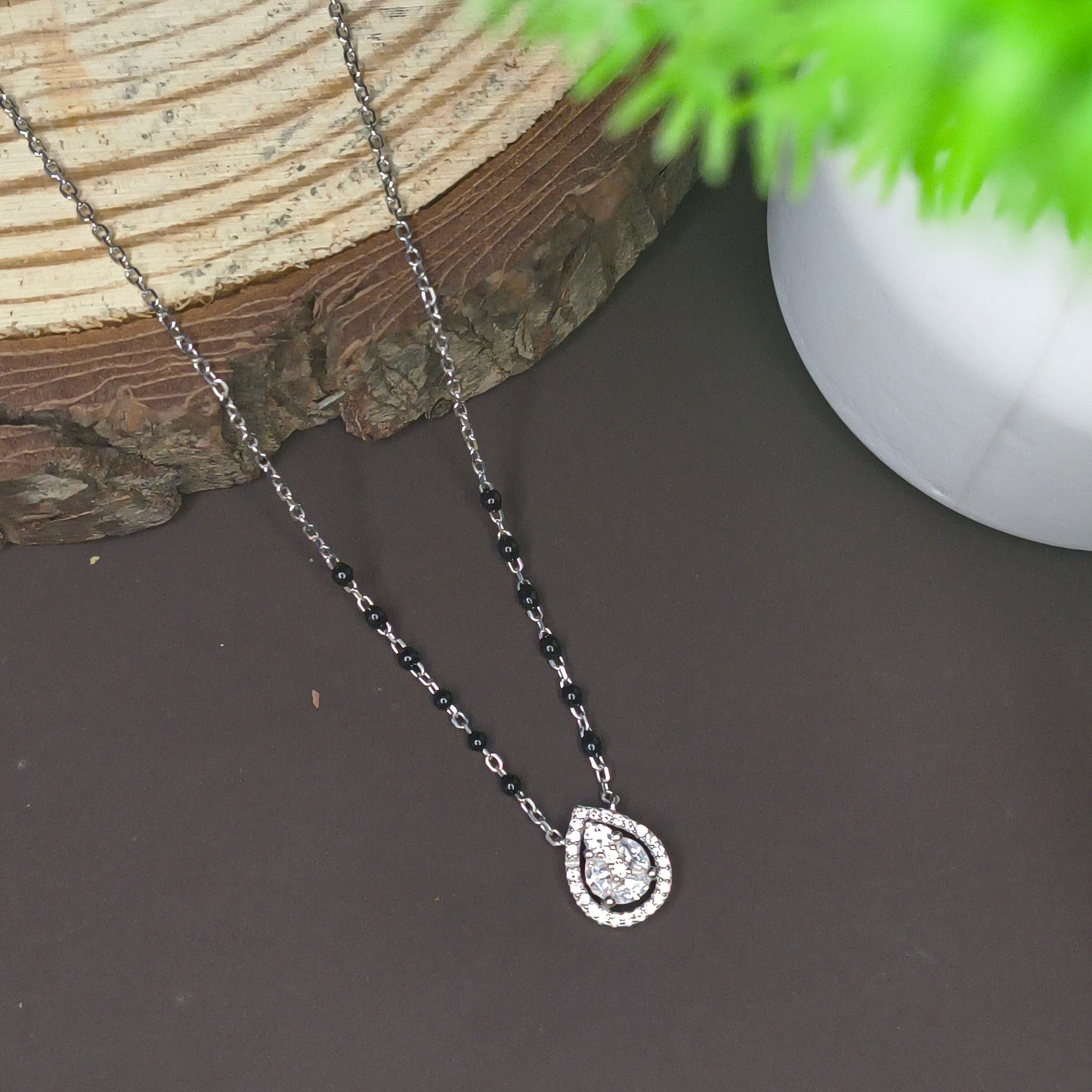 Jewelry with a teardrop-shaped pendant on a dark surface with a plant in the corner.