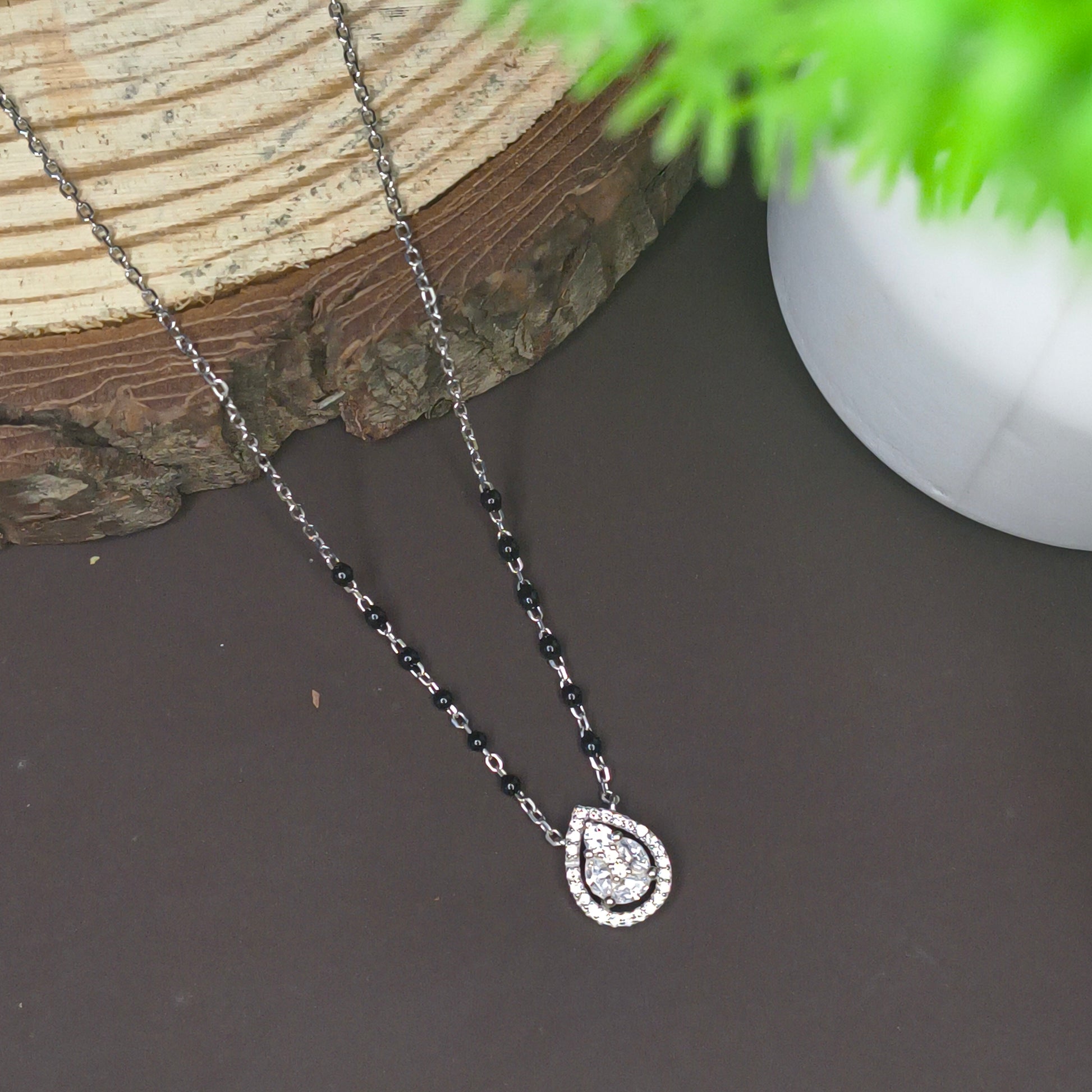 Jewelry with a teardrop-shaped pendant on a dark surface with a plant in the corner.