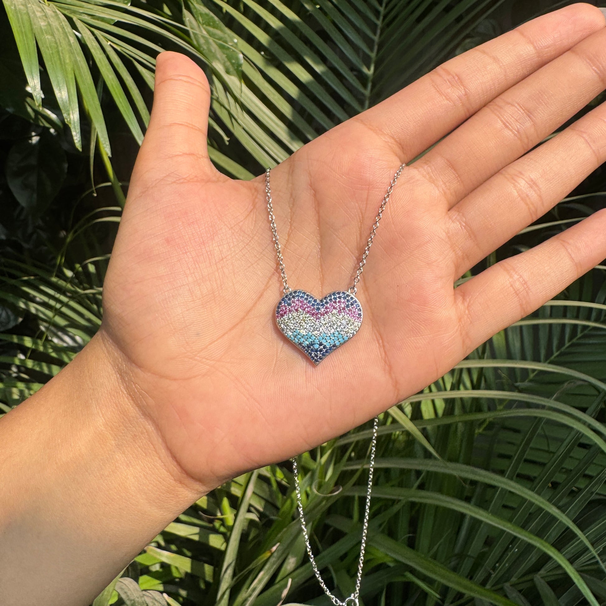 Hand holding a heart-shaped multicolored necklace against a green leafy background