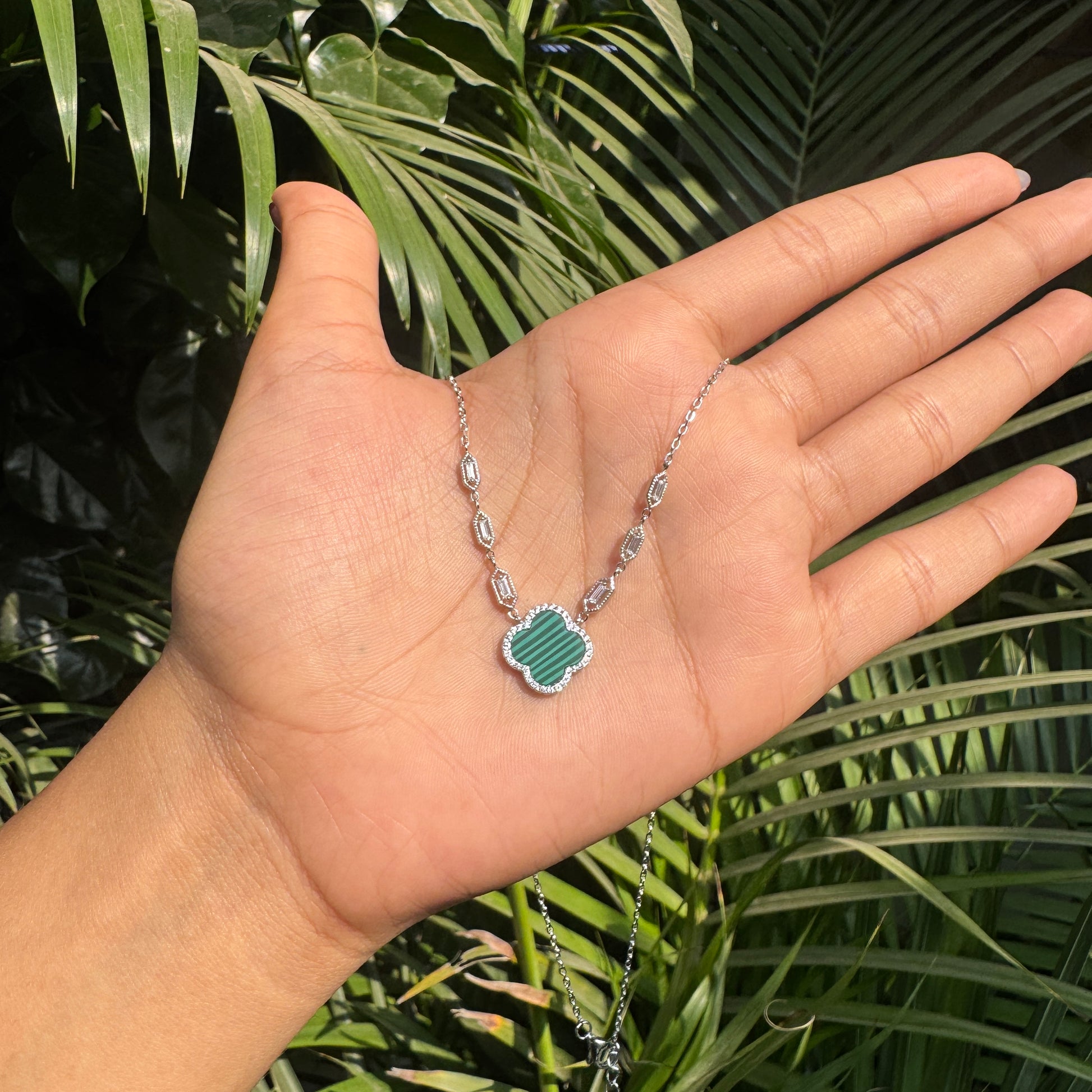 Hand wearing a silver necklace with a green pendant against a green leafy background