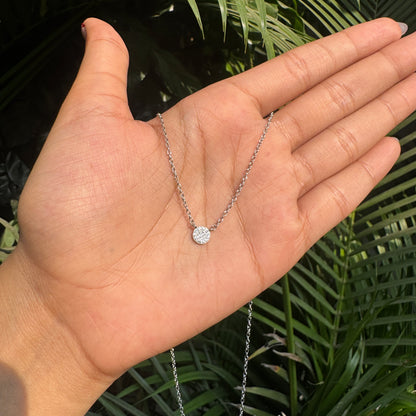 Hand holding a delicate silver necklace with a small round pendant against a green leafy background