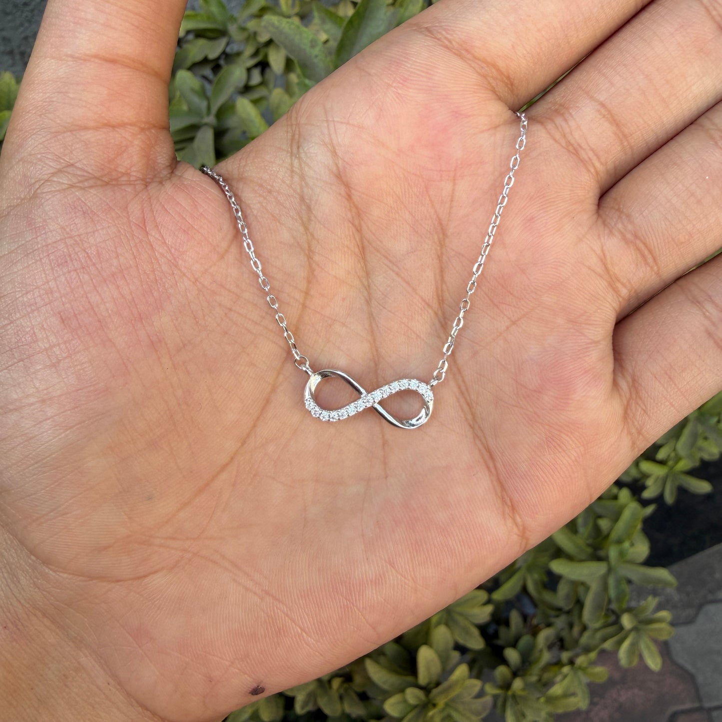 Silver infinity necklace on a hand with a green plant background