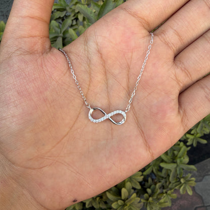 Silver infinity necklace on a hand with a green plant background