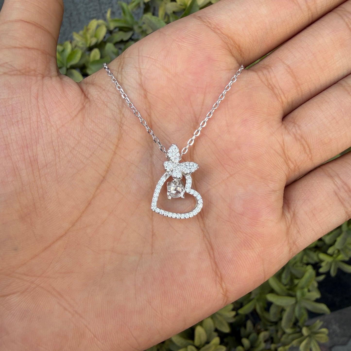 Silver necklace with a heart and angel pendant held in a hand against a natural background
