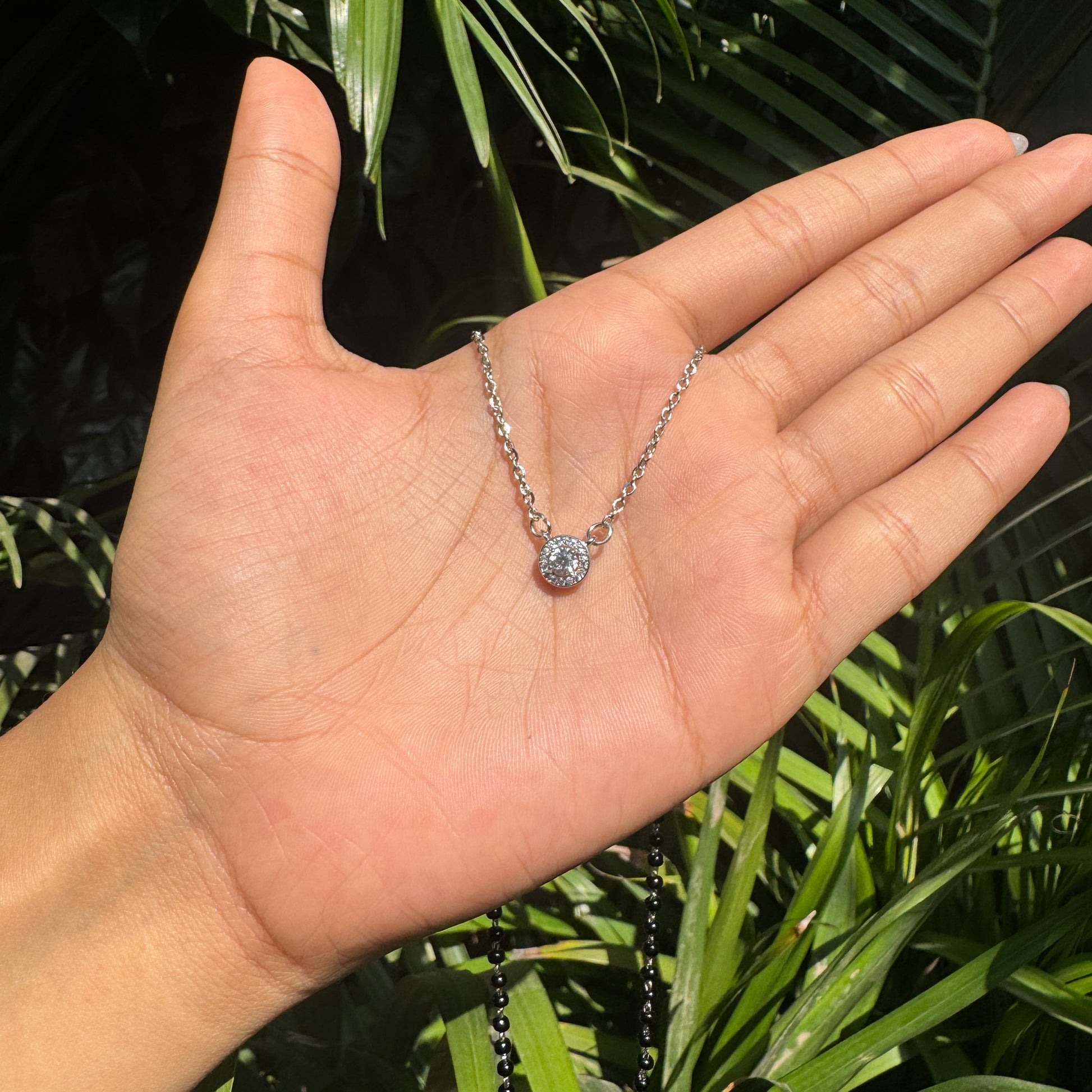 Hand holding a silver necklace with a clear pendant against a green leafy background