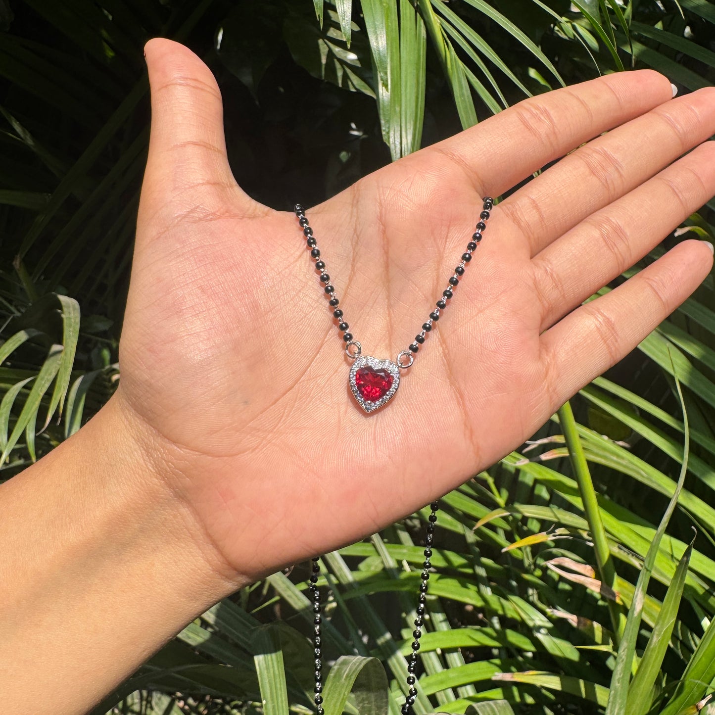 Hand holding a necklace with a heart-shaped red pendant against a green leafy background