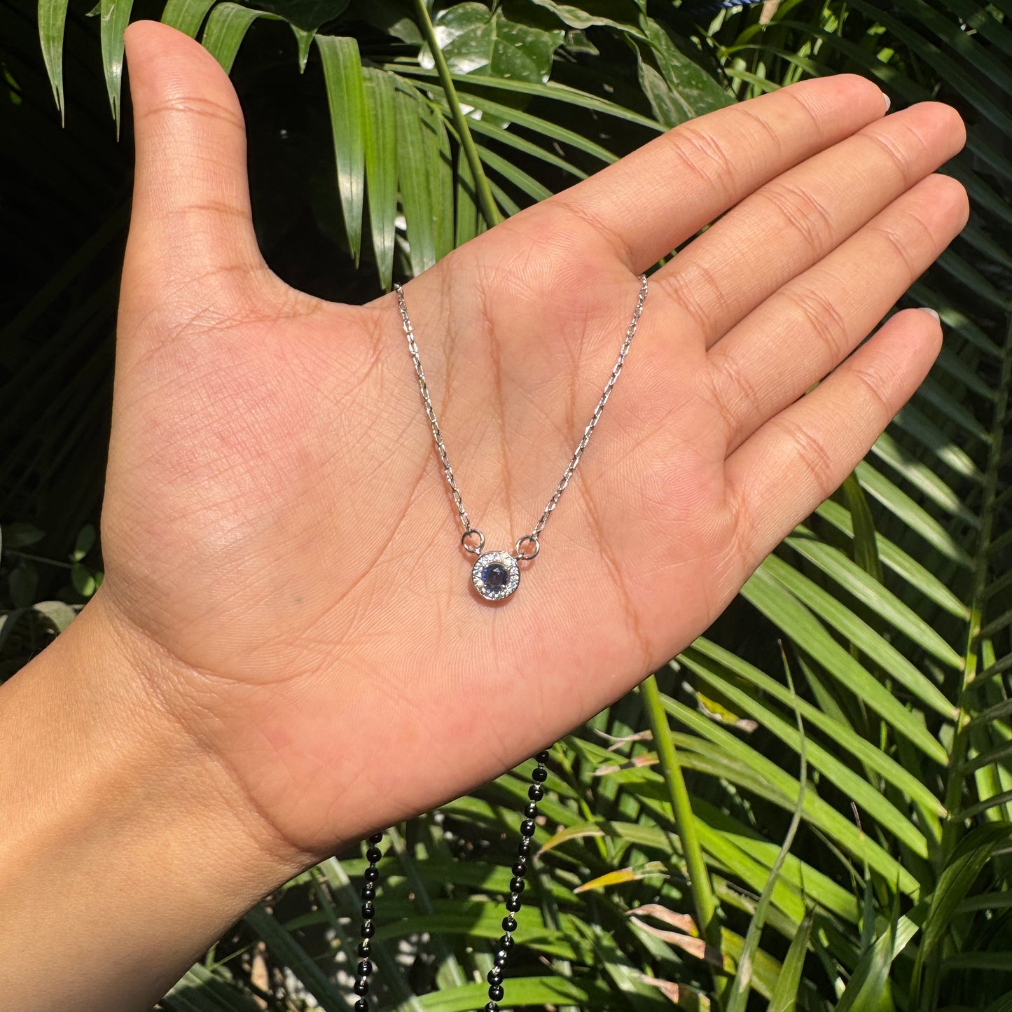 Hand holding a silver necklace with a blue gemstone against a green leafy background