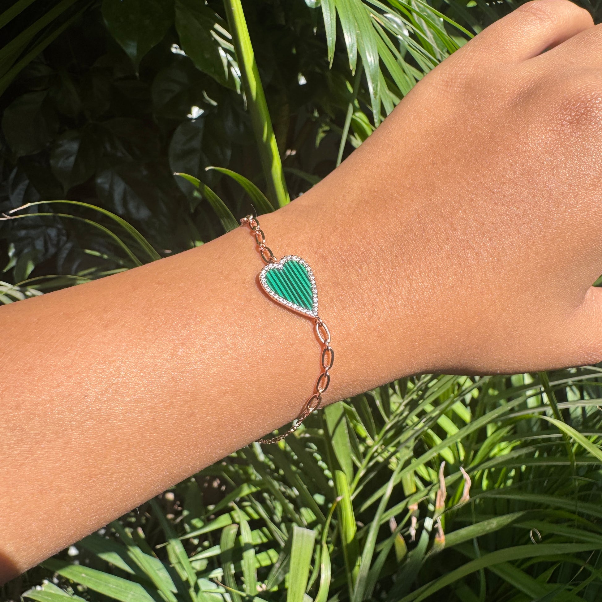 Hand wearing a green heart-shaped bracelet with a leaf design against a green leafy background
