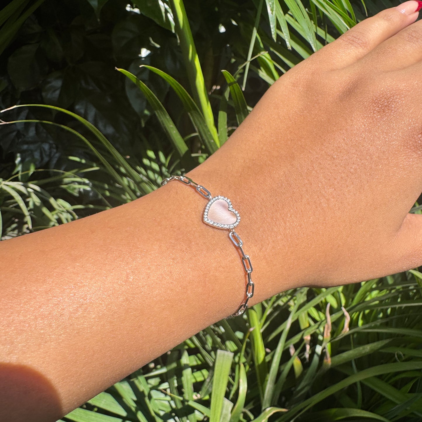 Hand wearing a silver bracelet with a heart-shaped charm against a green leafy background