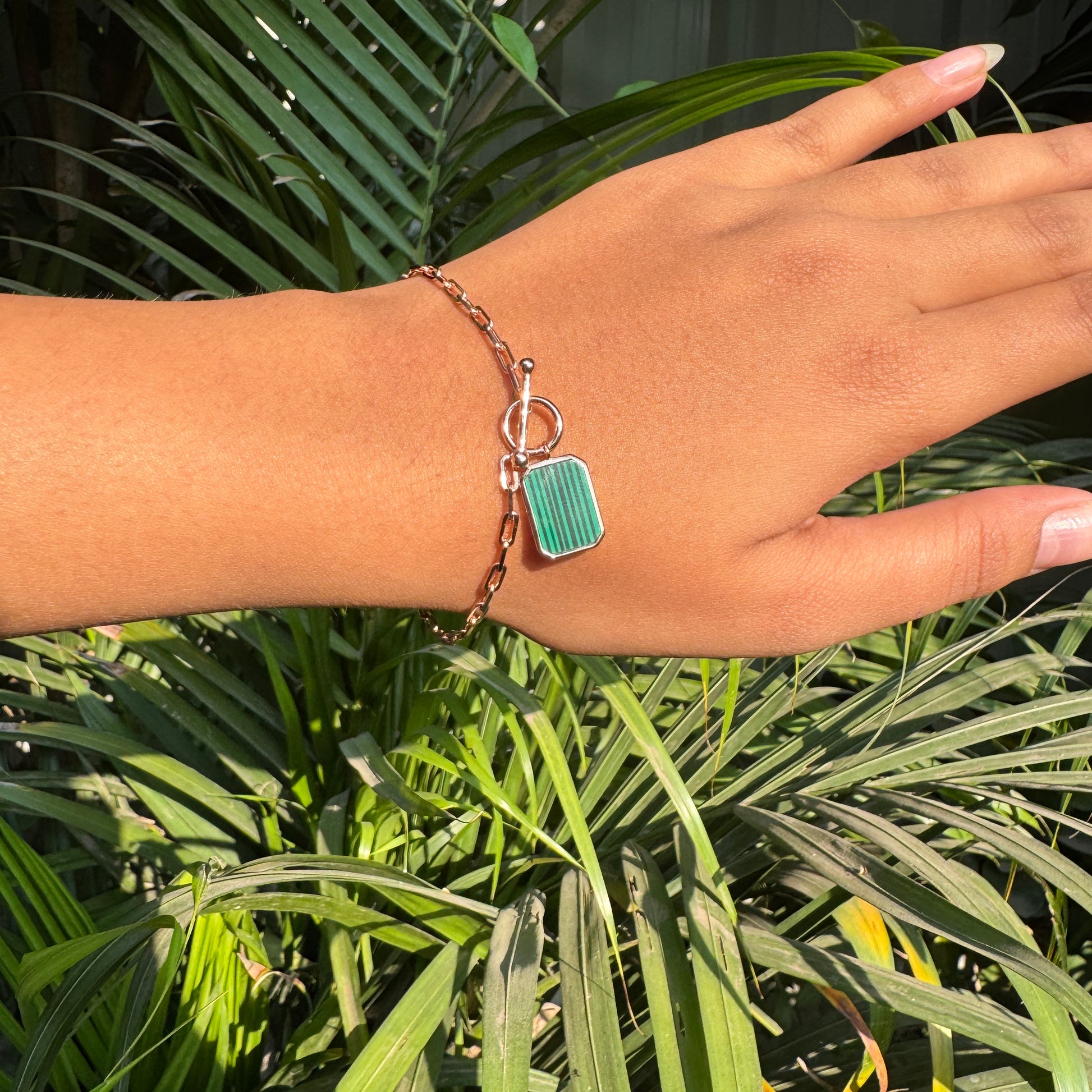 Hand wearing a bracelet with a green rectangular charm against a green leafy background