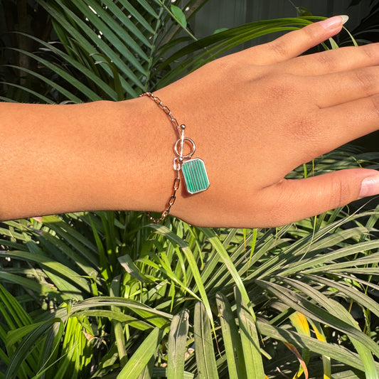 Hand wearing a bracelet with a green rectangular charm against a green leafy background