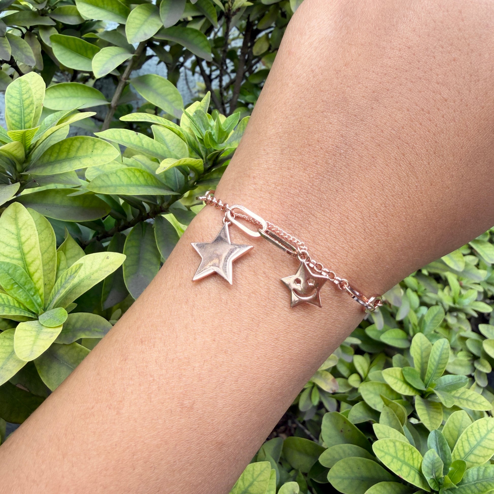 Rose gold bracelet with star charms on a wrist against a green leafy background