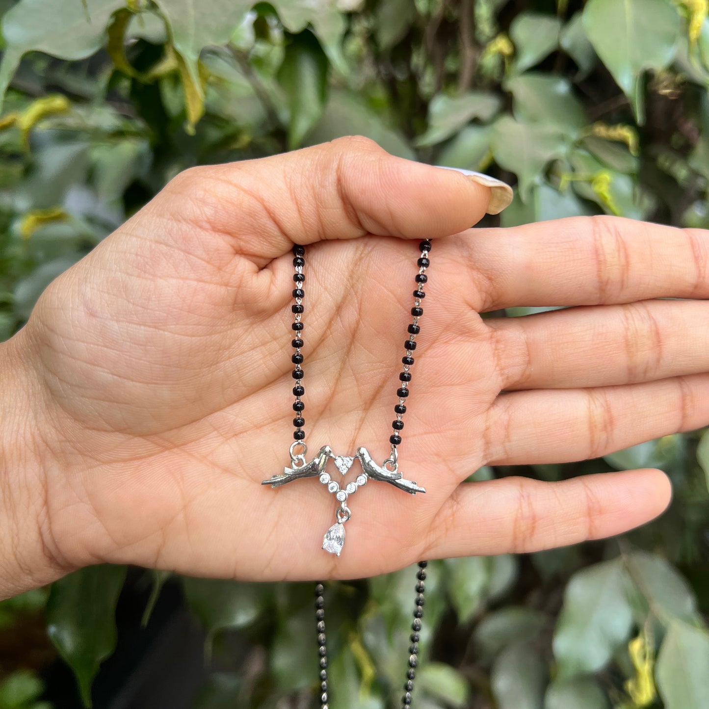 Hand holding a necklace with a silver pendant against a green leafy background