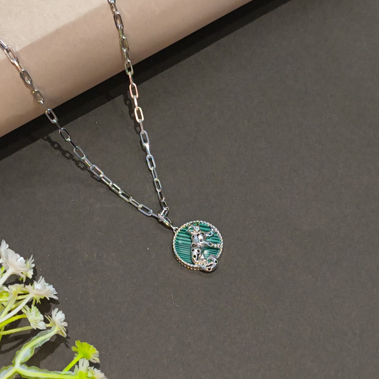 Necklace with a green pendant on a gray surface with a beige box and white flowers.
