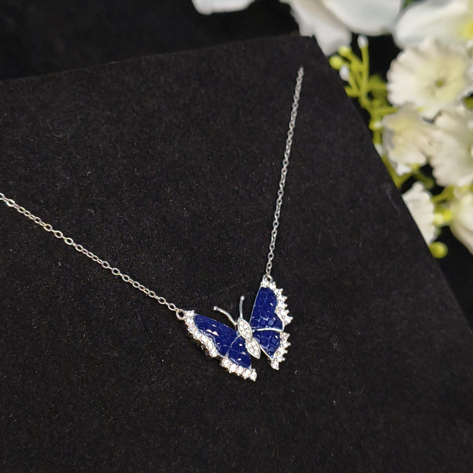 Butterfly-shaped necklace with blue and silver design on a black surface with flowers in the background