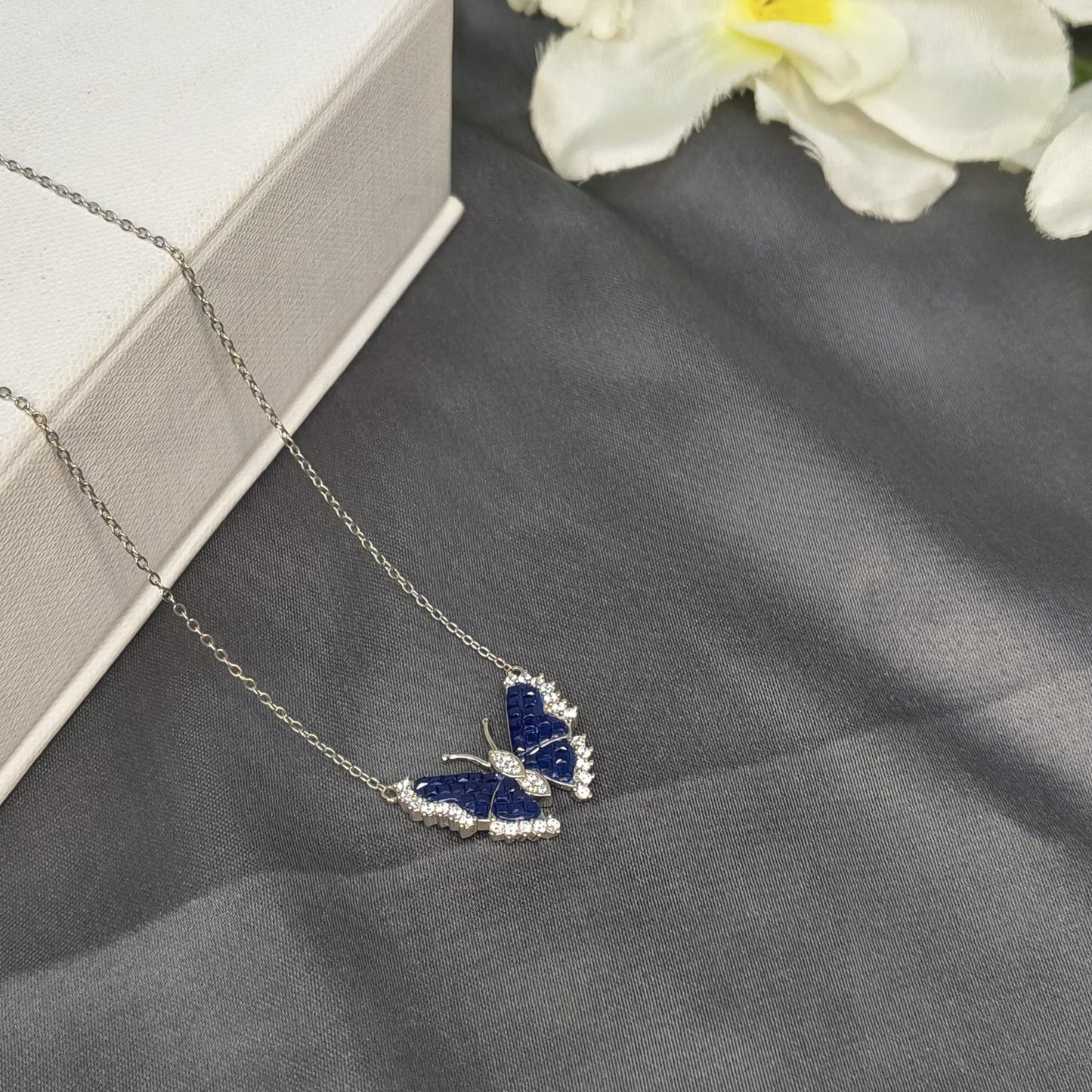 Butterfly-shaped necklace with blue stones on a gray surface with white flowers in the background