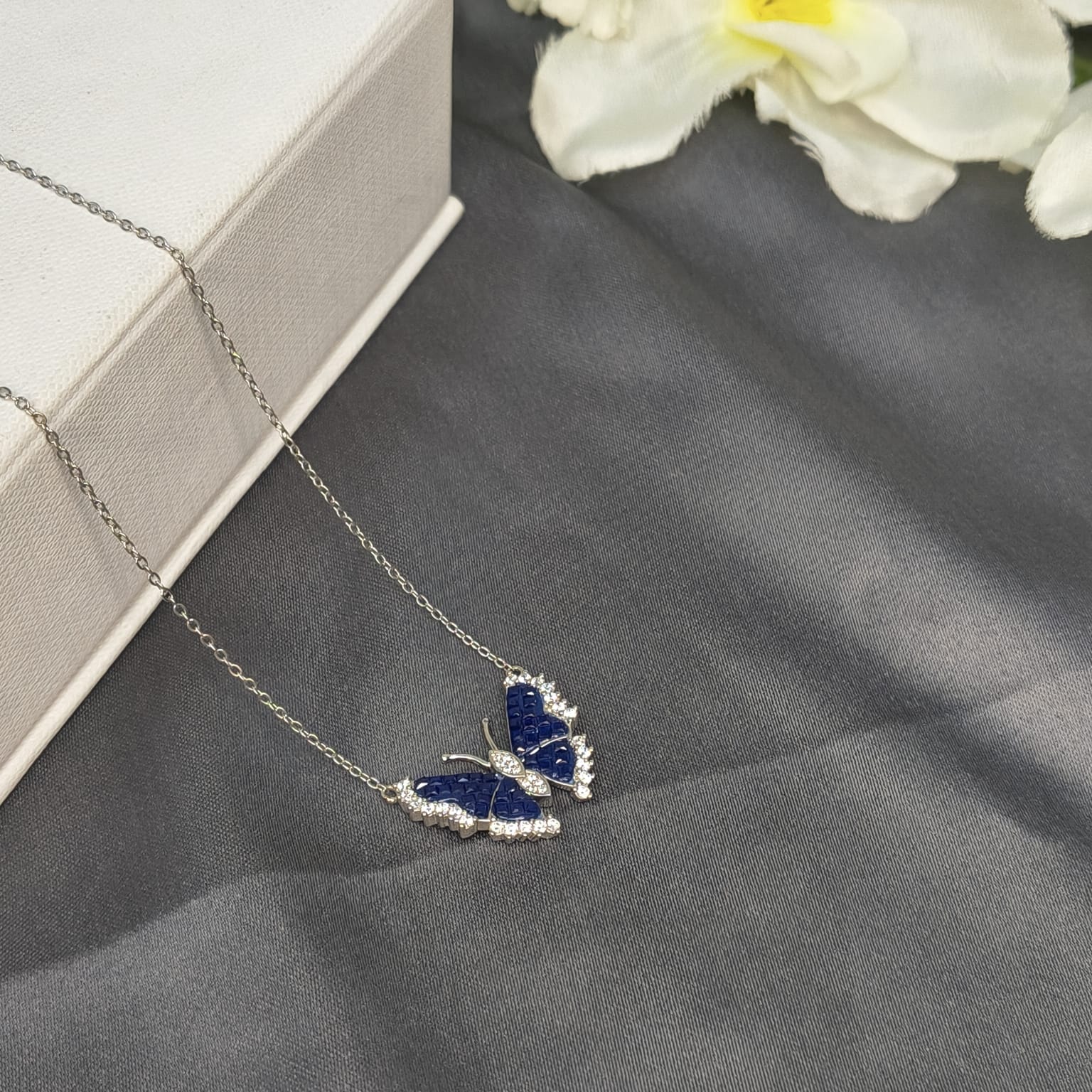 Butterfly-shaped necklace with blue stones on a gray surface with white flowers in the background