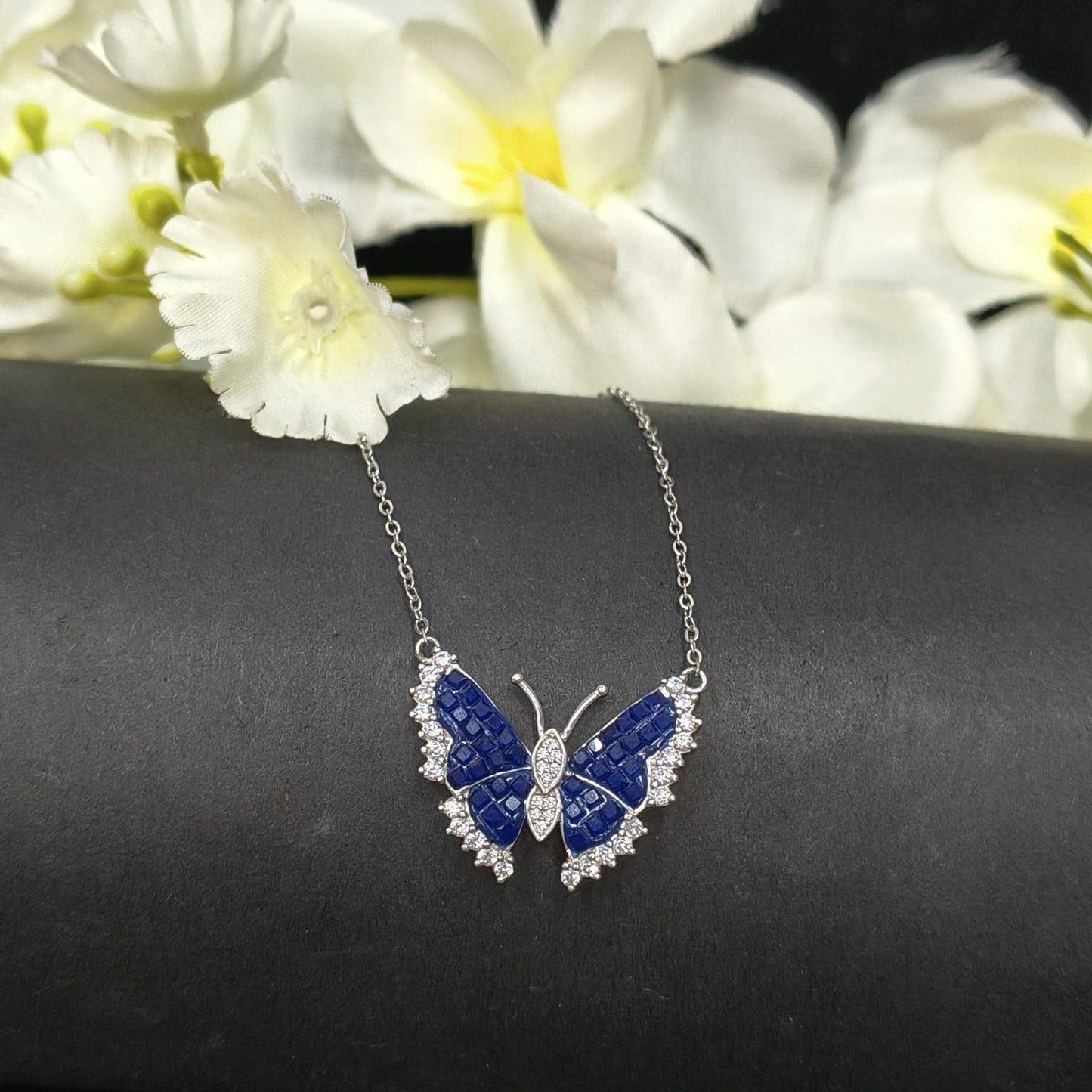 Butterfly-shaped necklace with blue and silver design on a dark surface with white flowers in the background