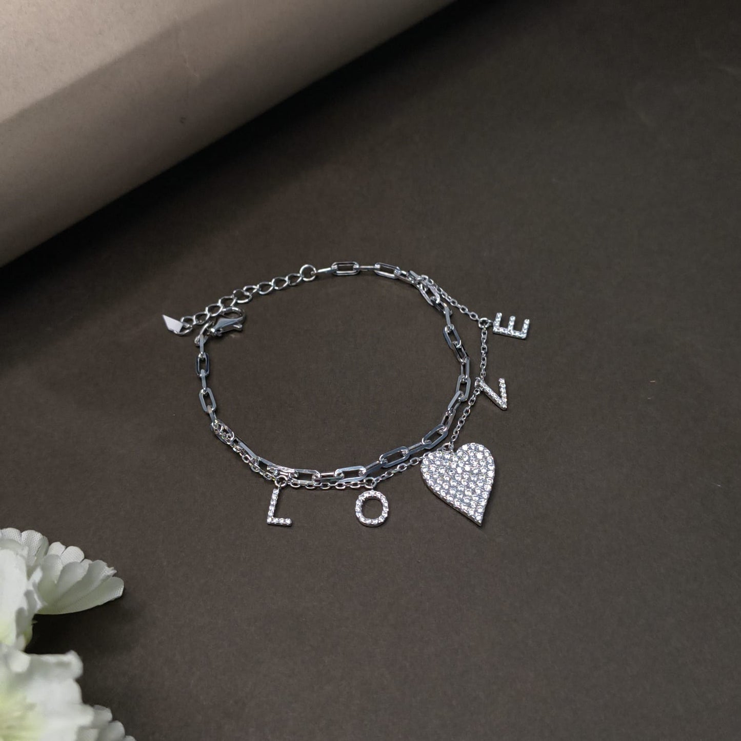 Silver bracelet with heart and letter charms on a dark surface with flowers.