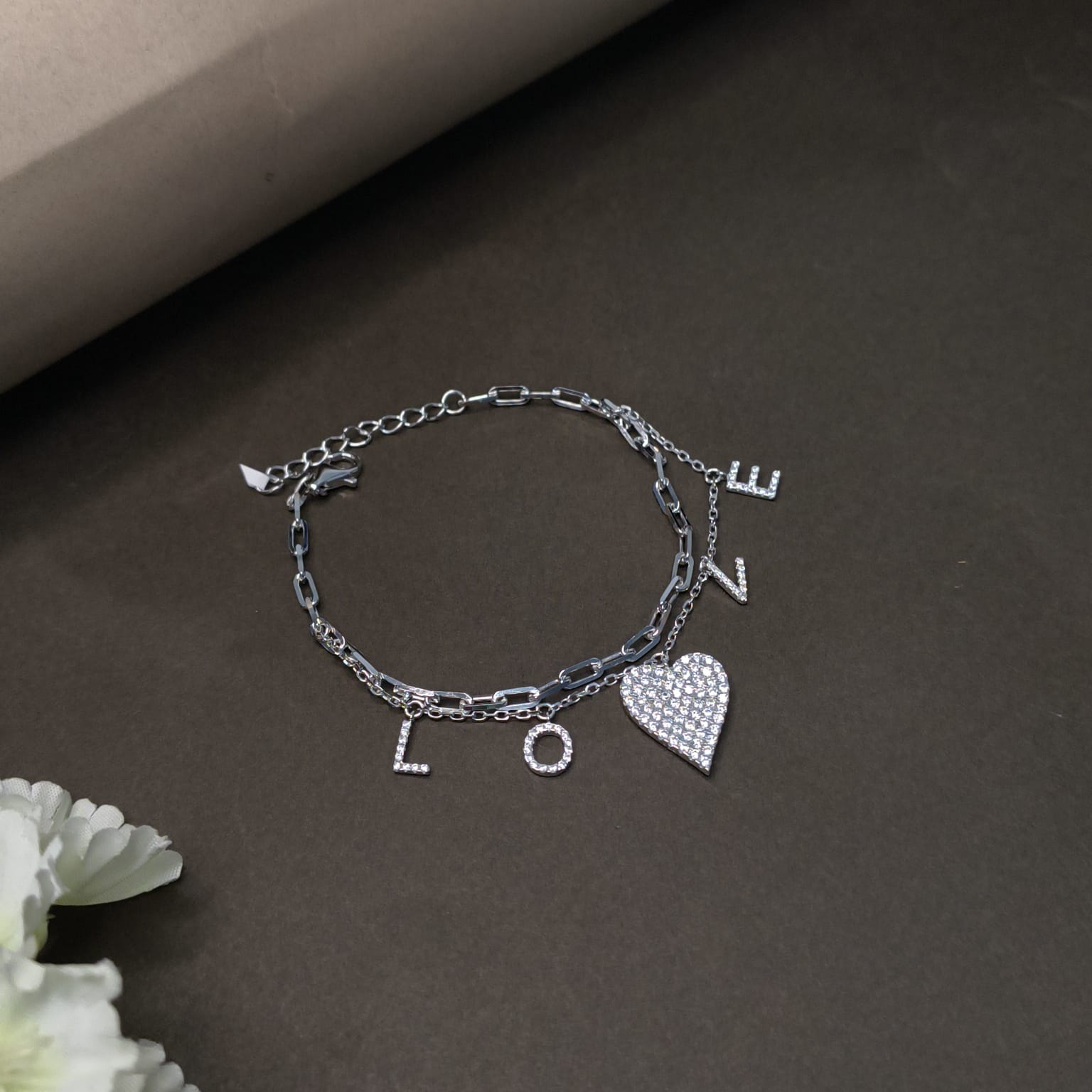 Silver bracelet with heart and letter charms on a dark surface with flowers.