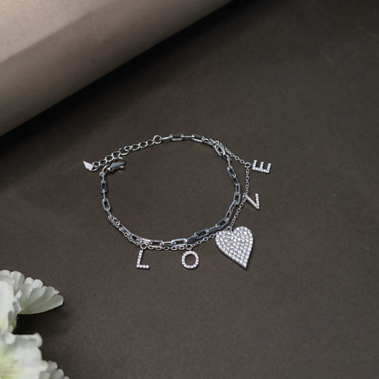 Silver bracelet with heart and letter charms on a dark surface with flowers.