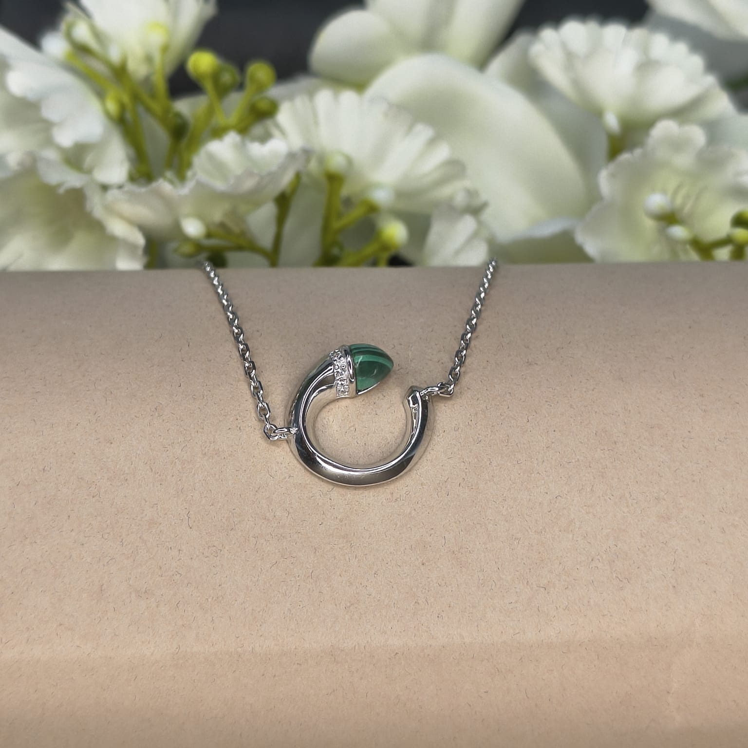 Silver necklace with a green pendant on a beige surface with white flowers in the background