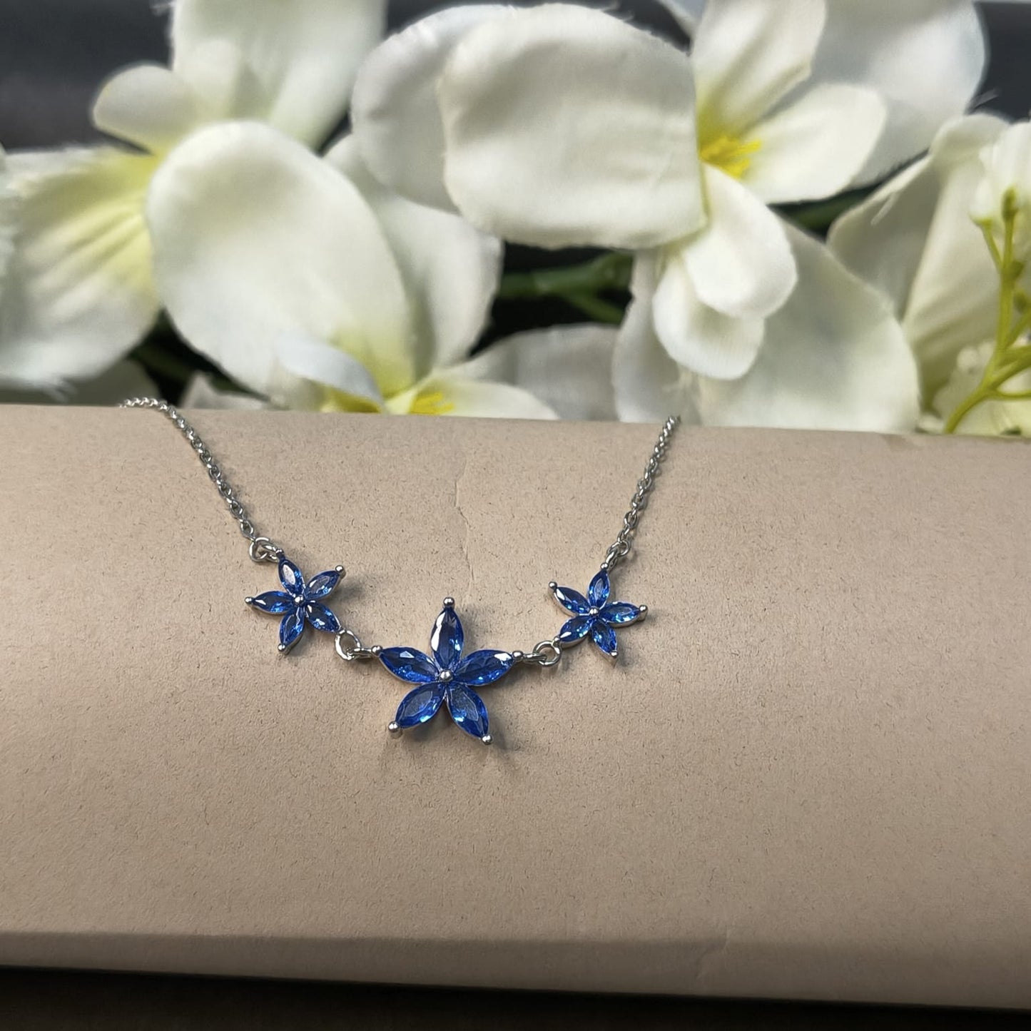 Necklace with blue flower-shaped pendants on a beige surface with white flowers in the background