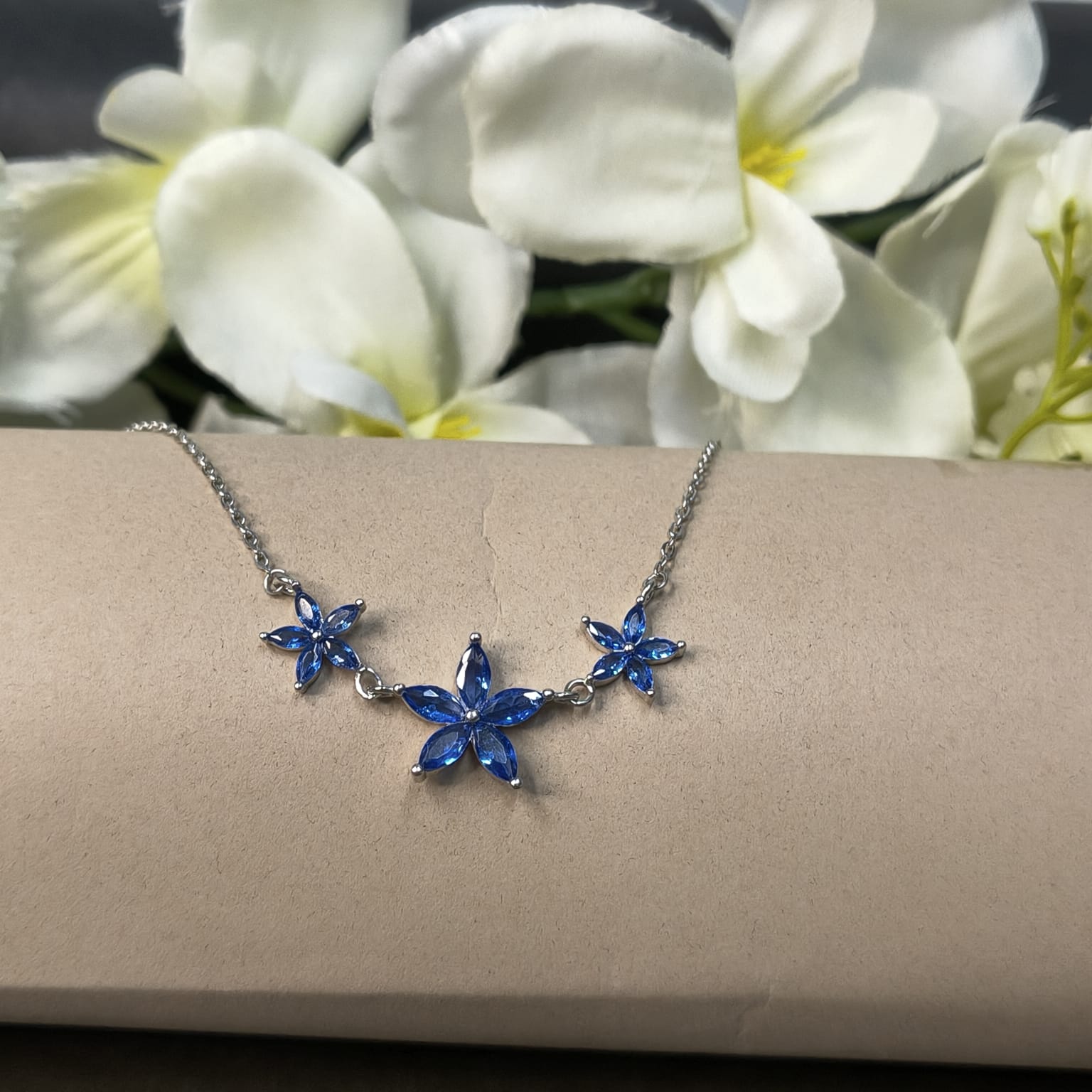 Necklace with blue flower-shaped pendants on a beige surface with white flowers in the background