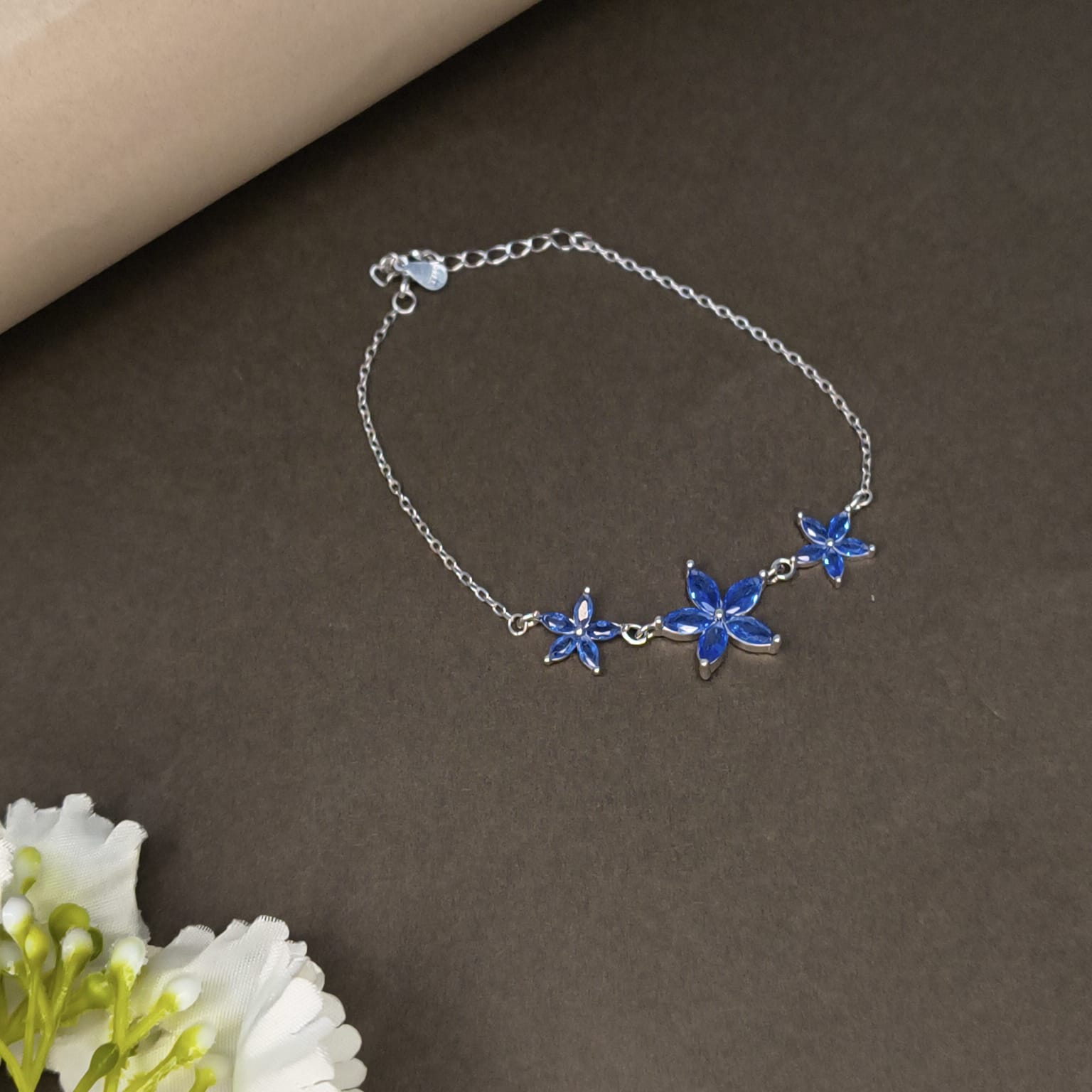 Silver bracelet with blue flower charms on a brown surface with white flowers.
