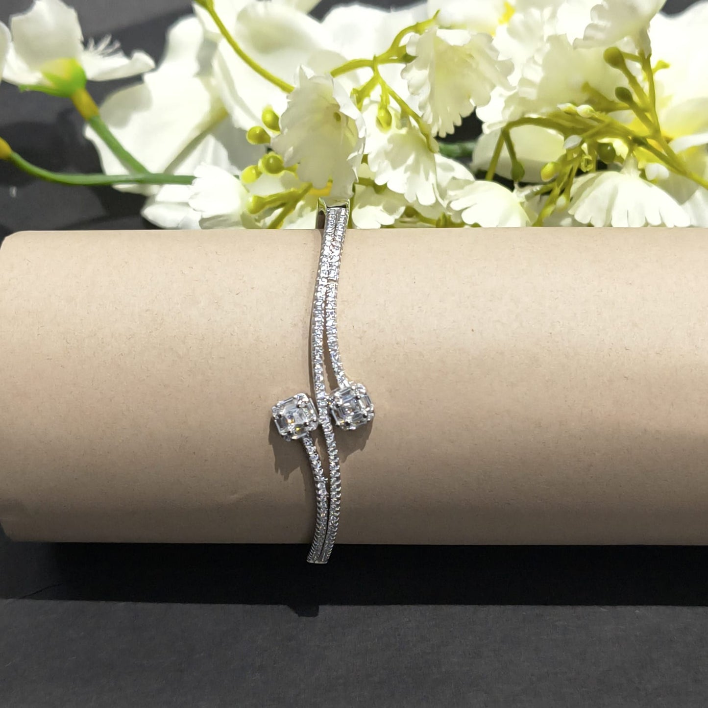 Silver bracelet with two clear stones on a cylindrical cardboard roll with white flowers in the background