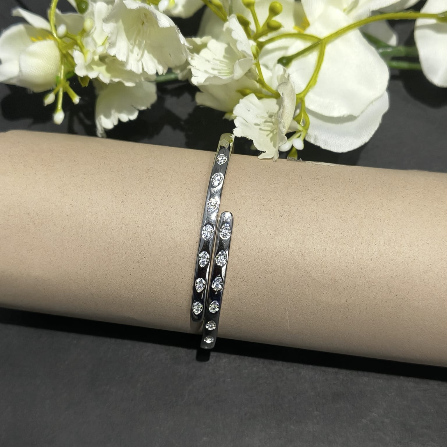 Silver bracelet with clear stones on a beige cylindrical object with white flowers in the background