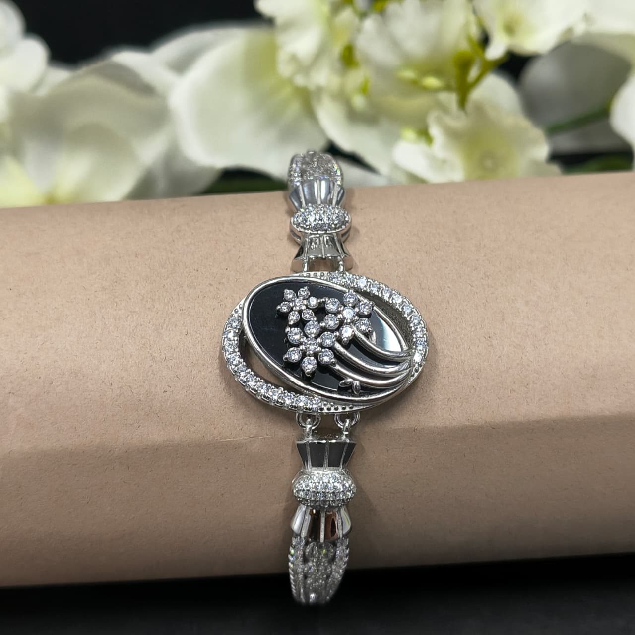 Silver bracelet with black center and floral design on a wrist, with white flowers in the background.