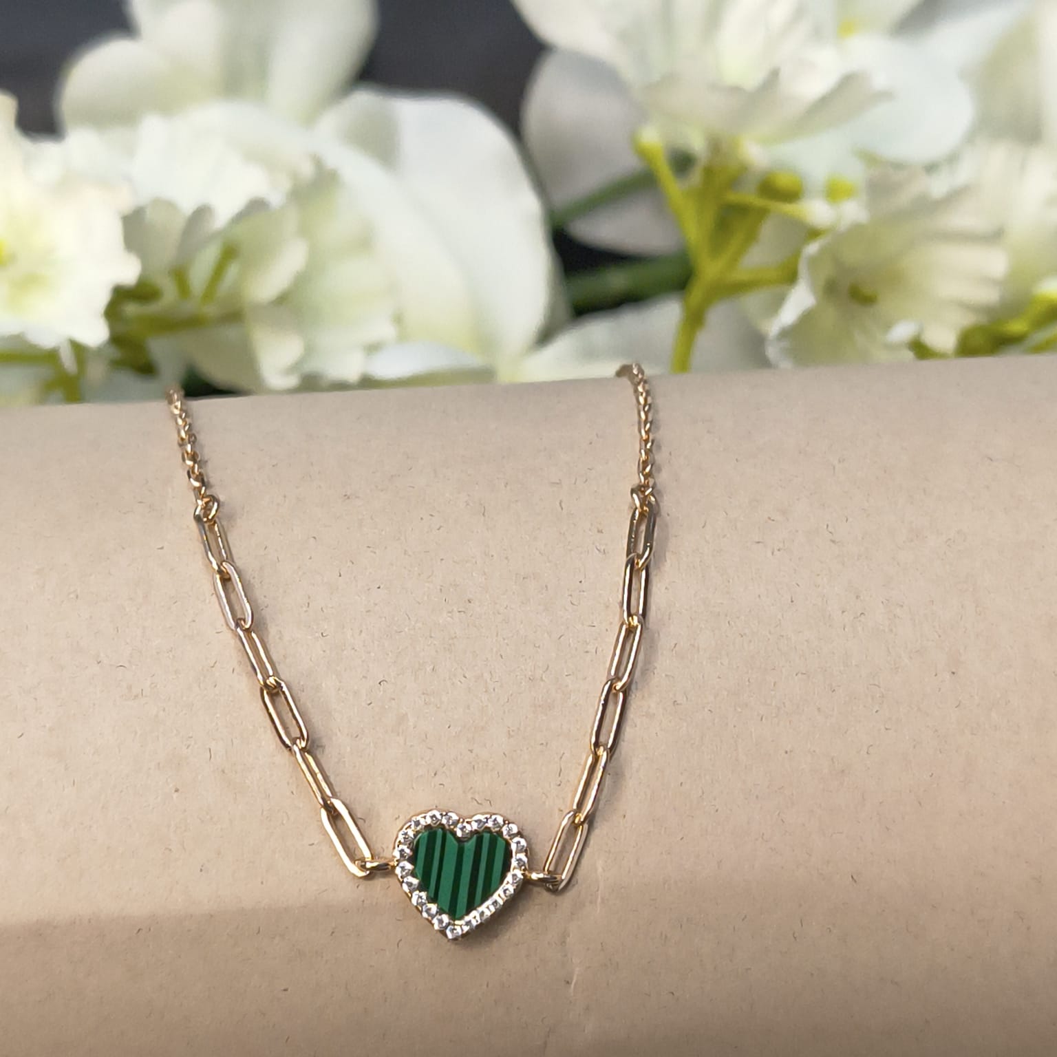 Gold chain necklace with a heart-shaped green stone on a wrist against a floral background