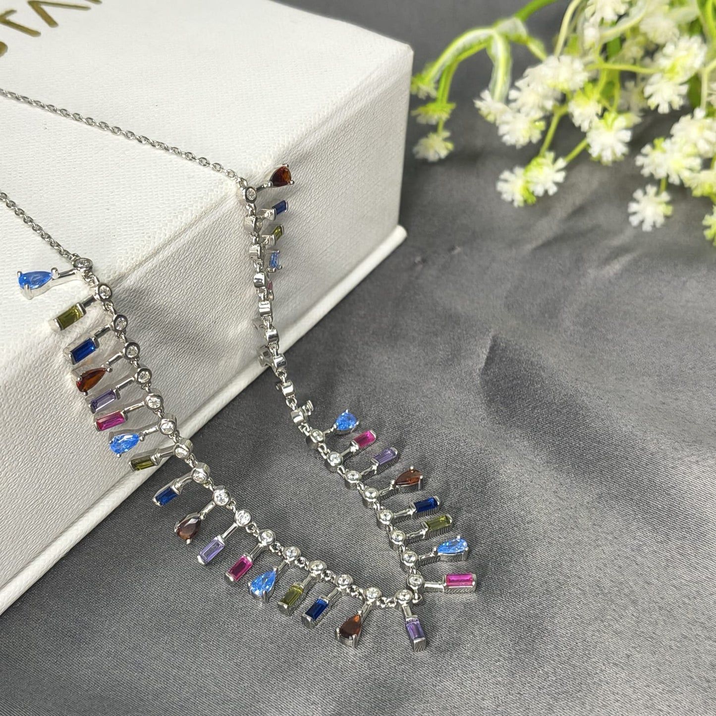 Necklace with colorful beads on a gray surface with a white box and flowers in the background
