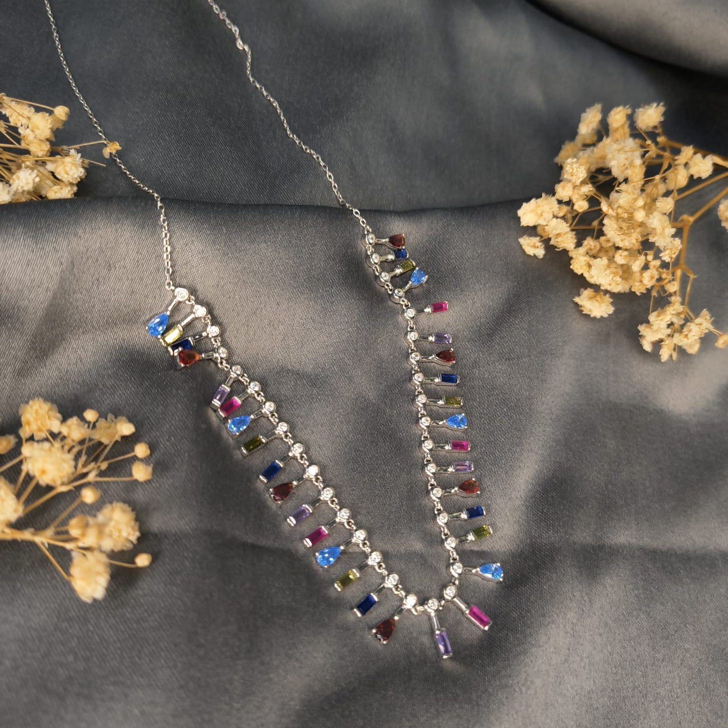 Necklace with multicolored gemstones on a gray fabric background with dried flowers.